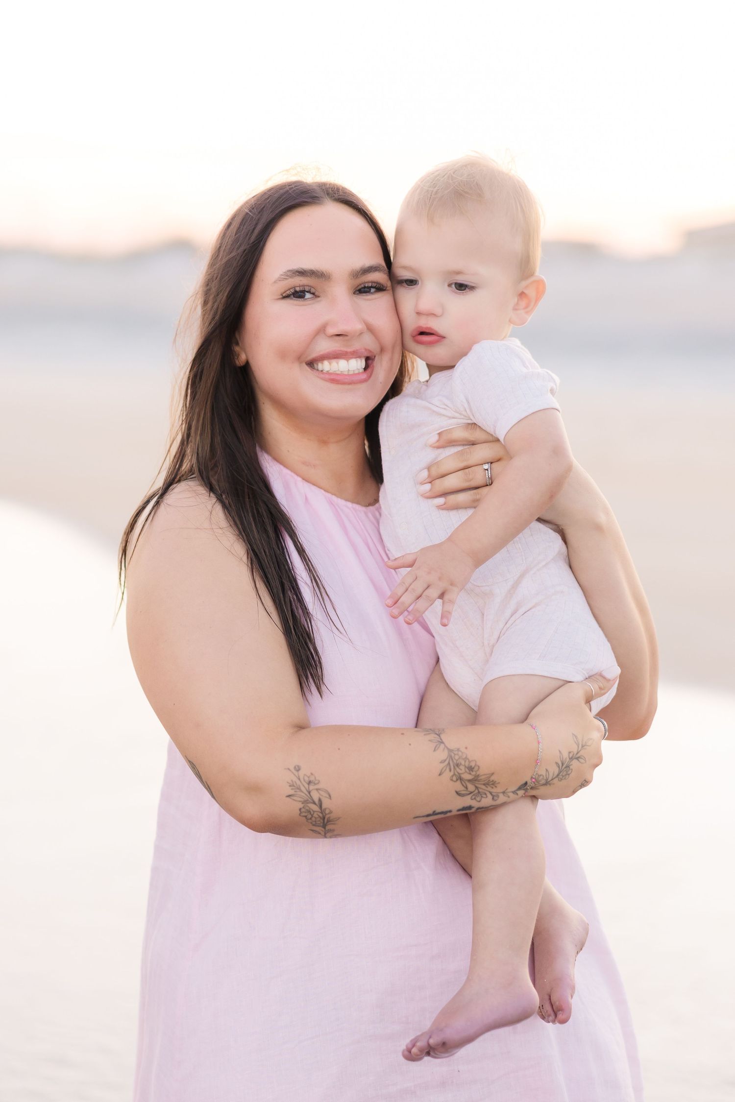 A smiling mother with dark hair and floral tattoos holds her baby on a serene sandy beach at sunset.