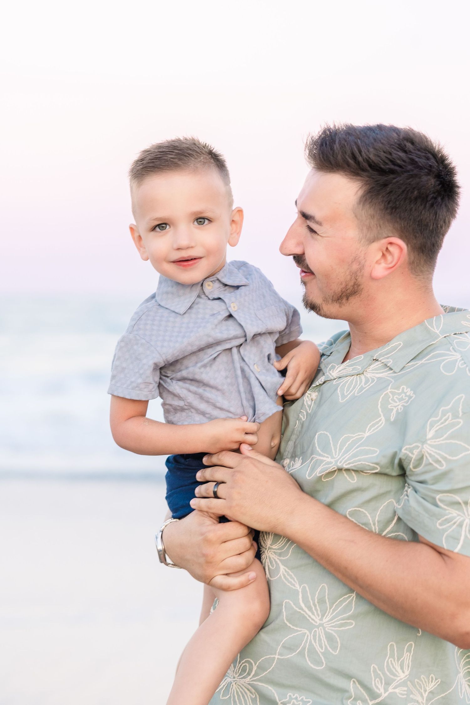 A father holding his young son on a beach with ocean waves in the background.