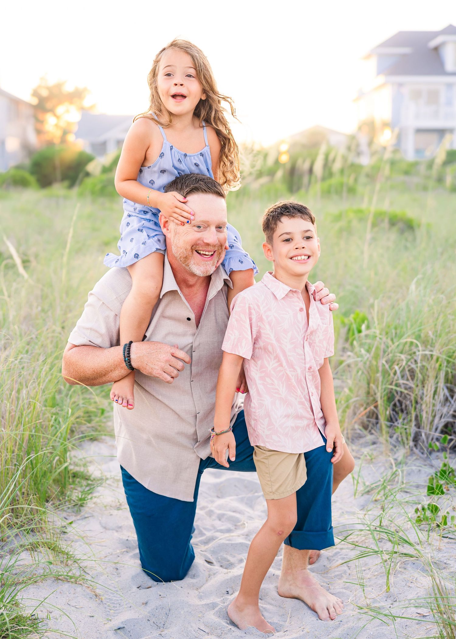 A father kneels on a sandy beach path with his daughter on his shoulders and son beside him.