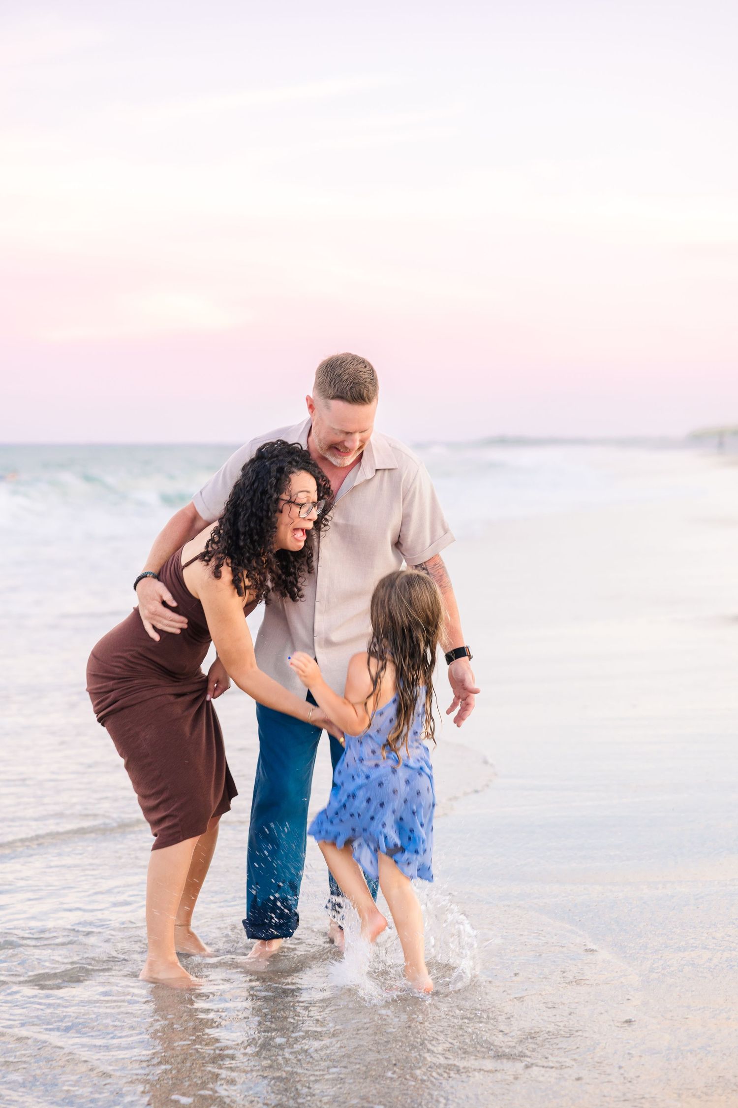 Parents laugh with young daughter splashing in ocean waves at sunset on a beautiful pink-hued beach.