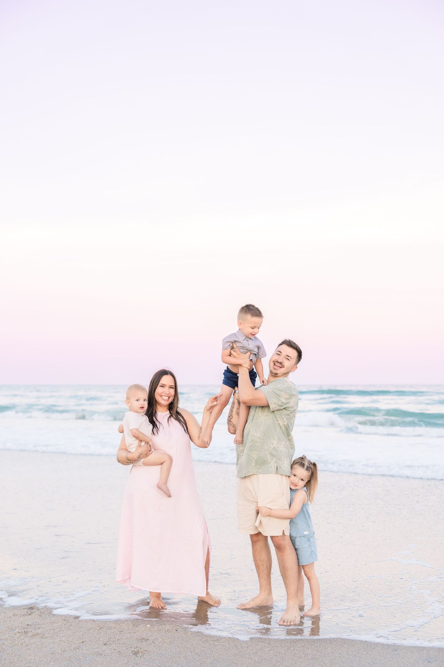 Joyful family beach portrait at dusk with parents lifting children and smiling near ocean waves.
