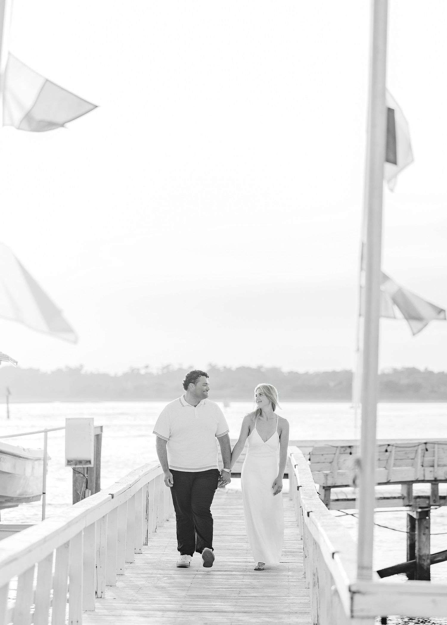 Black and white photo of a couple holding hands and walking on a dock with flags waving in the breeze.