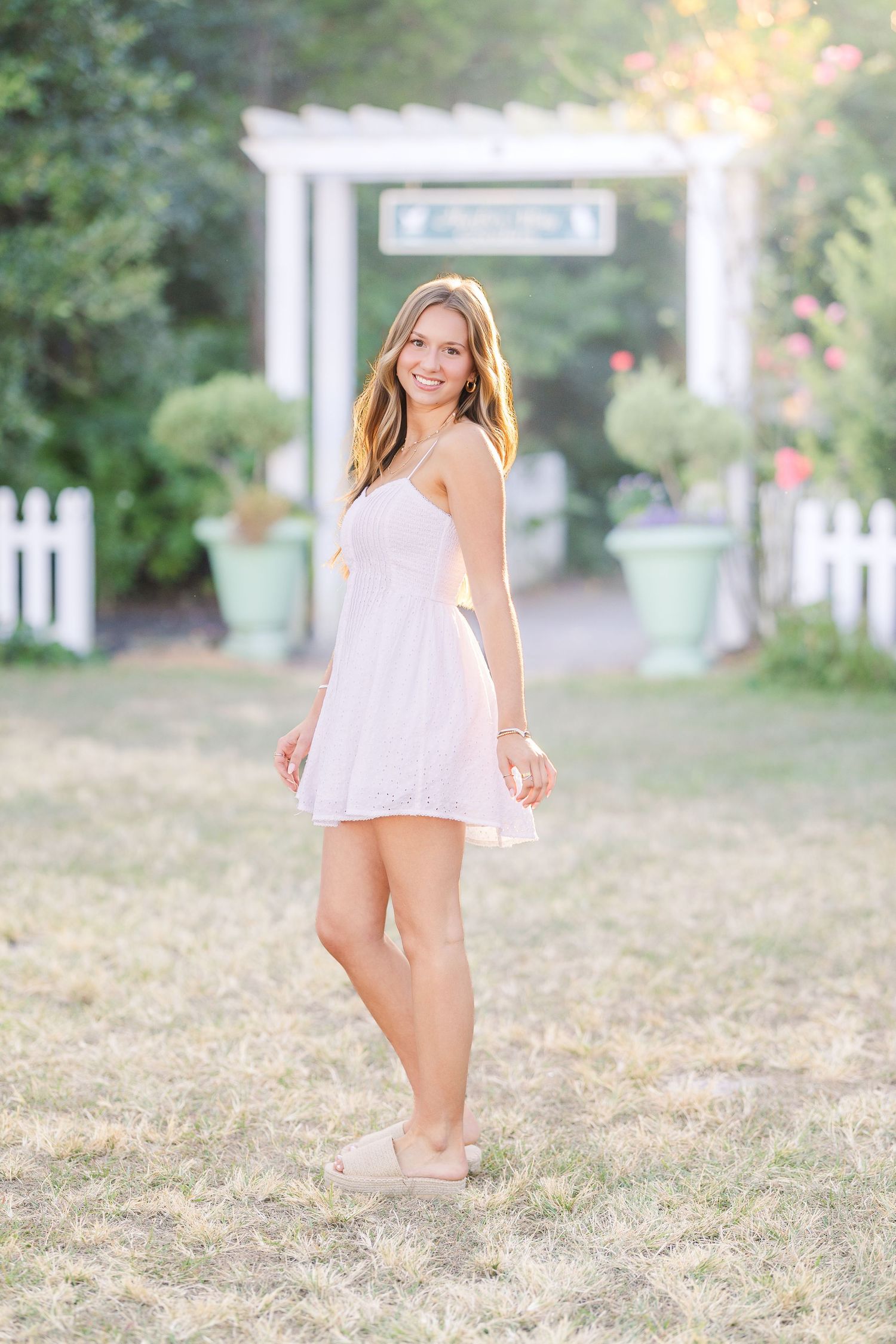 Person in white summer dress poses playfully near a white picket fence at sunset.