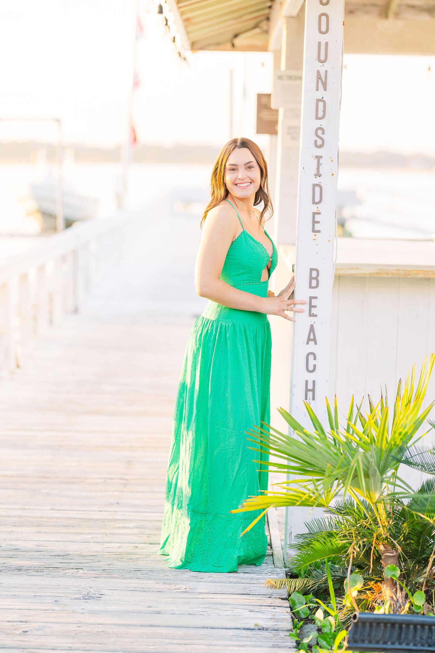 A flowing green maxi dress stands elegantly by a Soundside Beach sign on a wooden pier.