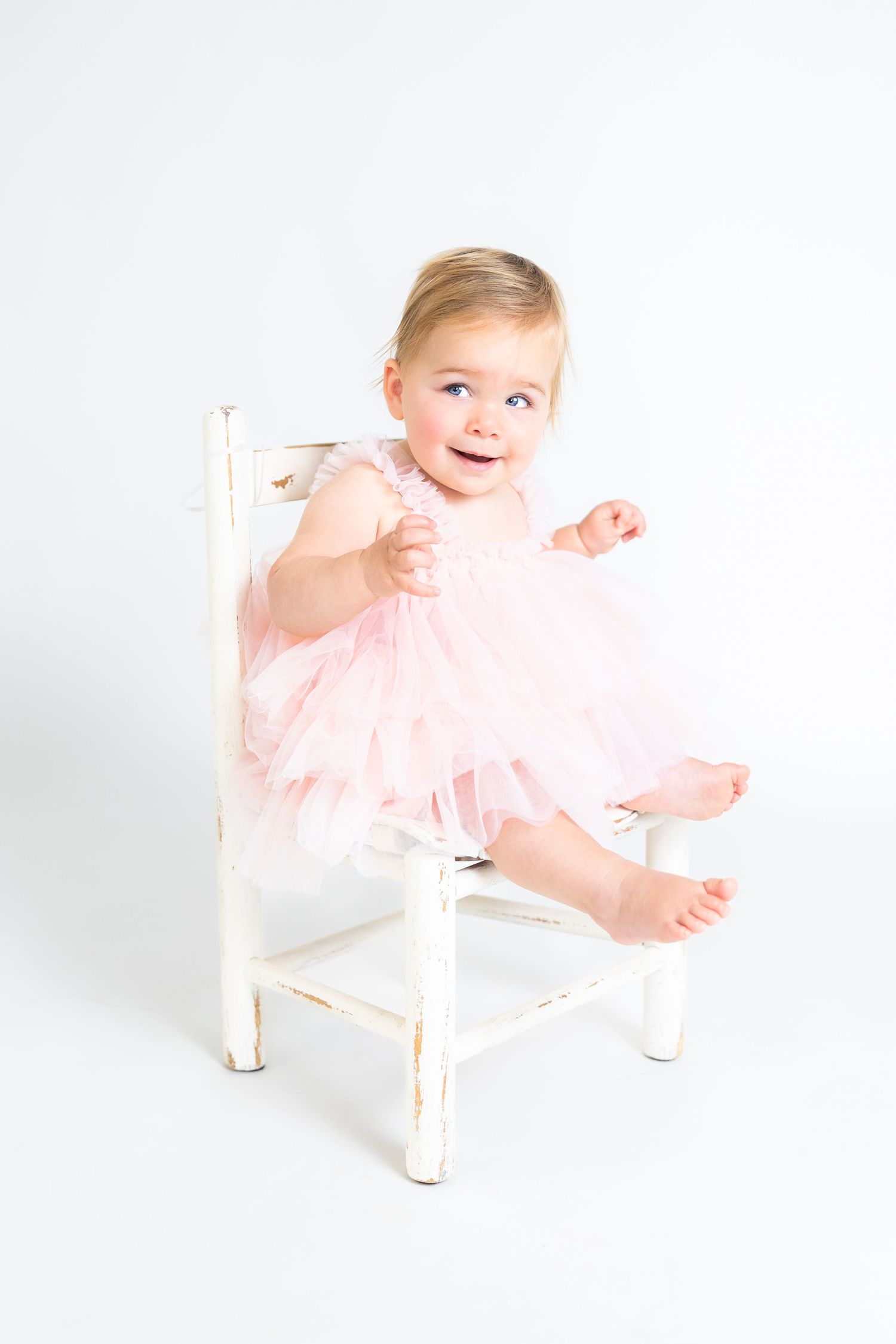 Baby wearing pink dress poses playfully on white chair against white background.