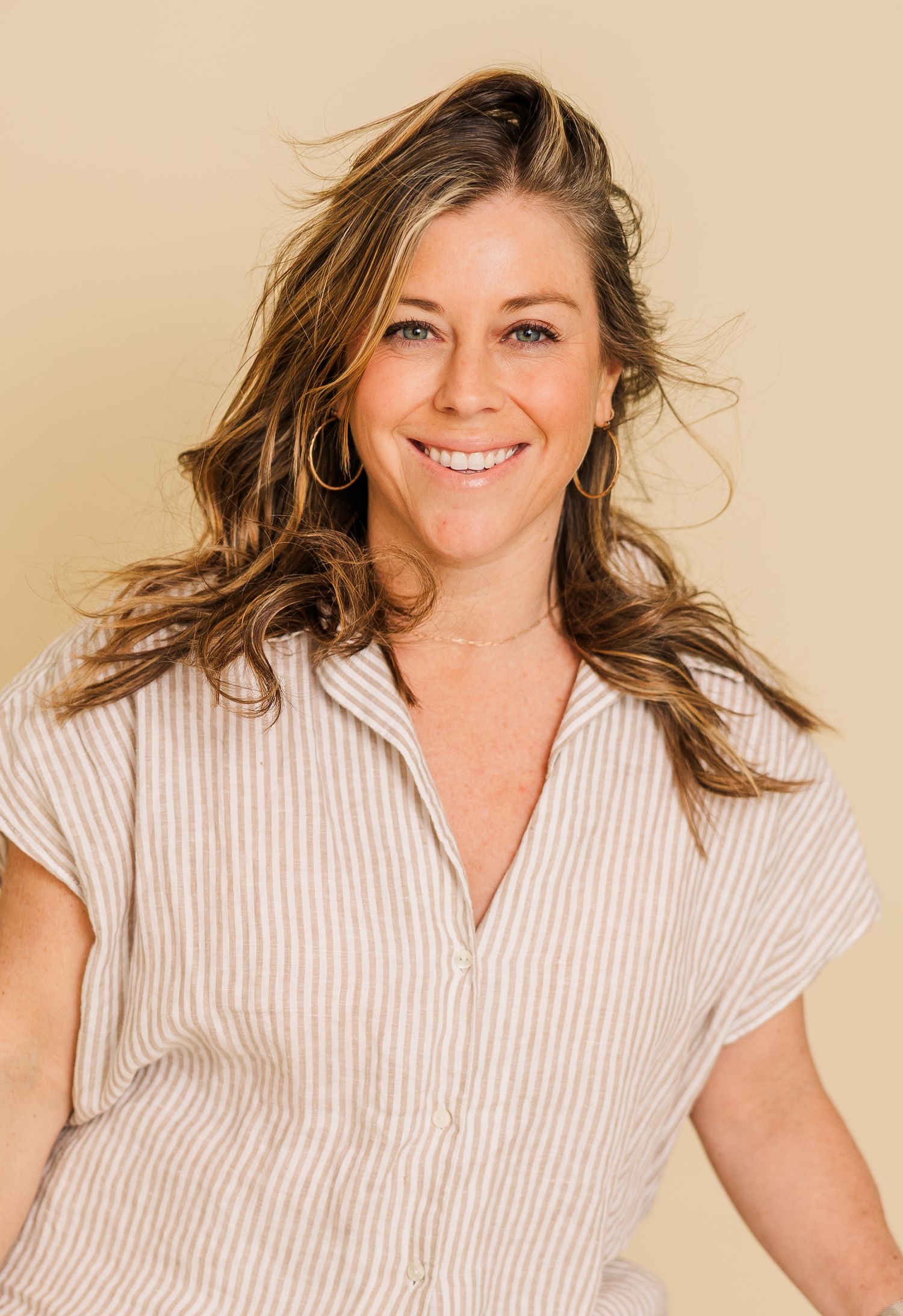 Woman in striped button-down shirt poses against beige background with windswept hair and natural smile.