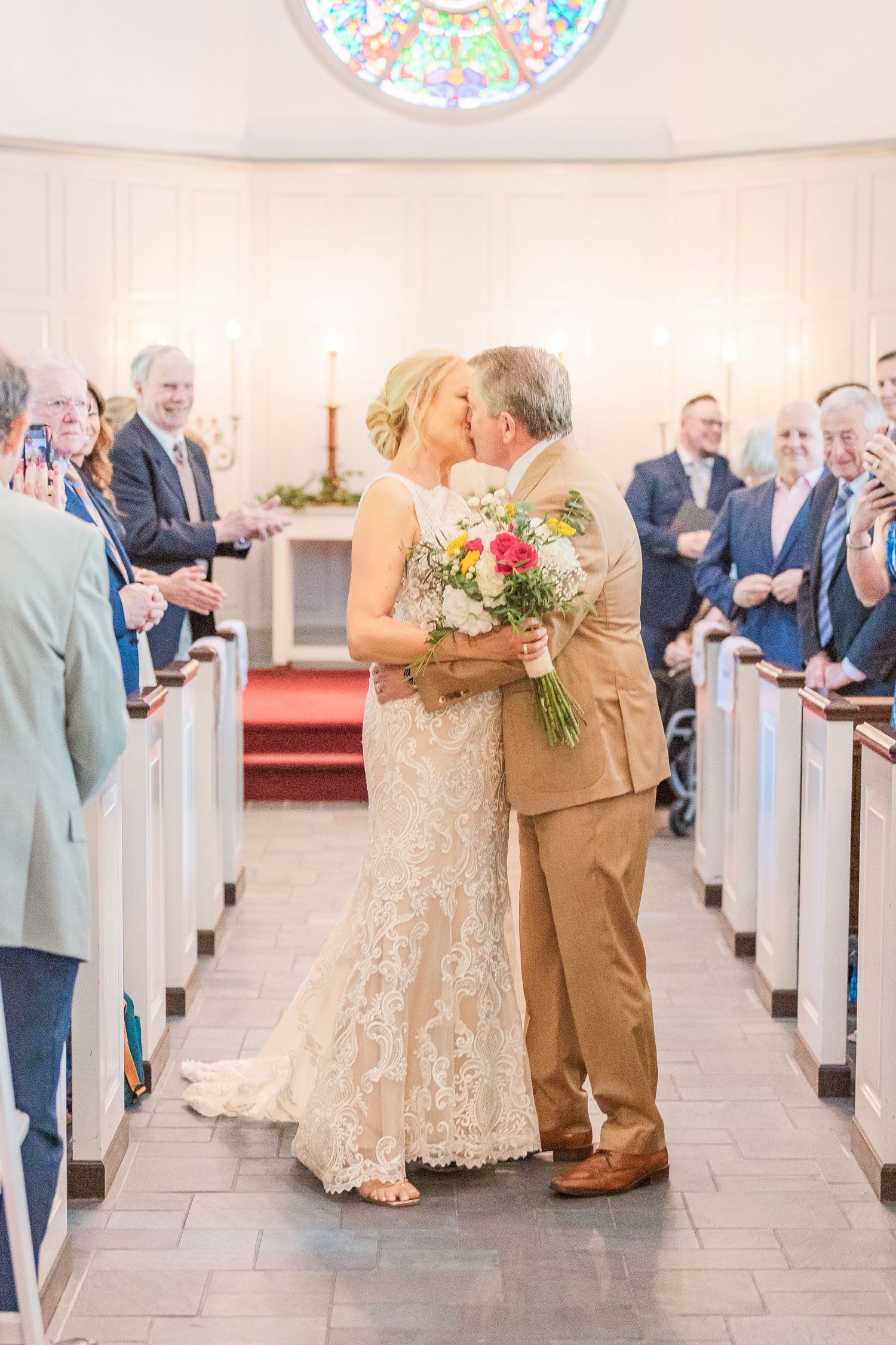 Wedding ceremony in a bright church with guests seated in white pews and sunlight streaming through stained glass windows.