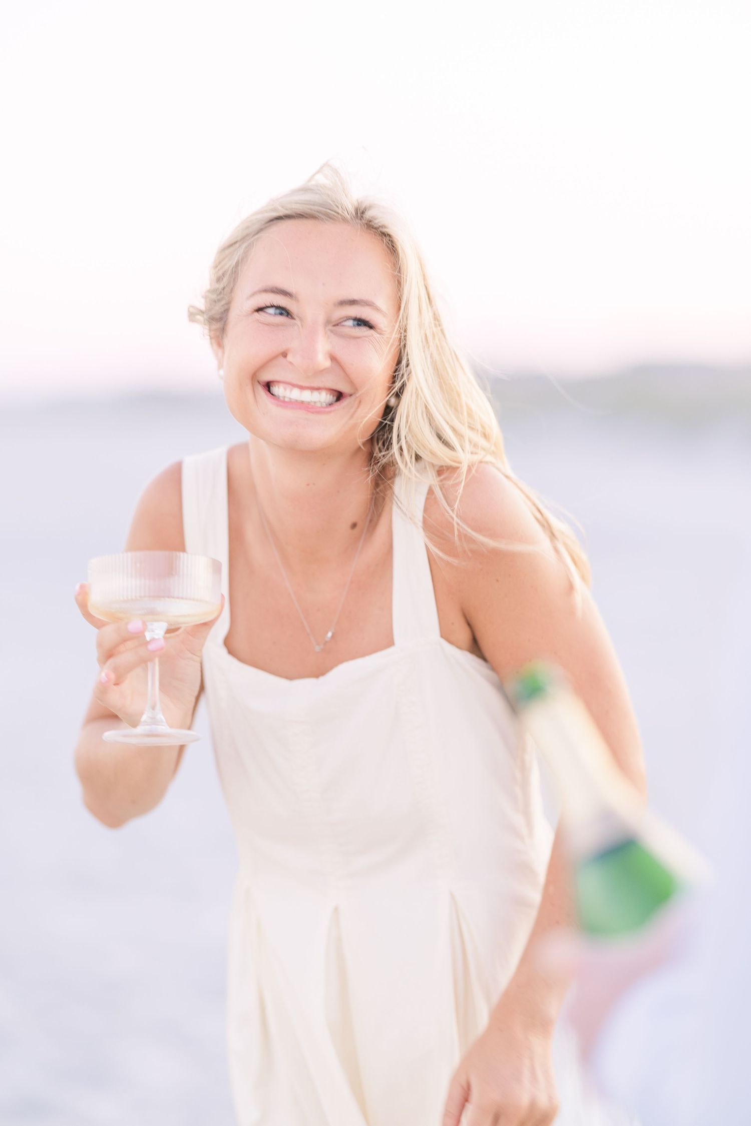 Person in white dress holding champagne and glasses on a bright beach setting.