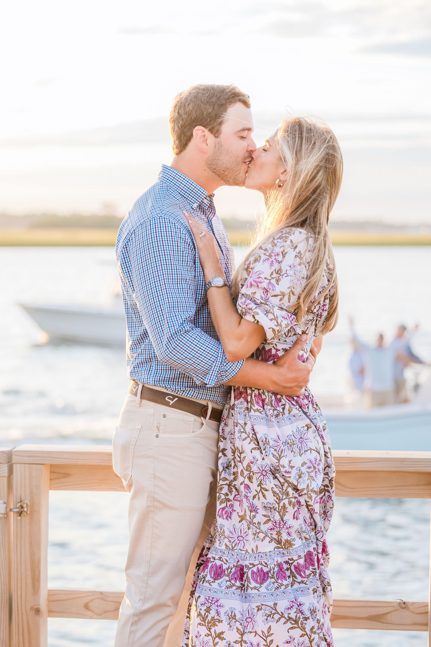 A romantic kiss shared between a couple on a waterfront deck at sunset.