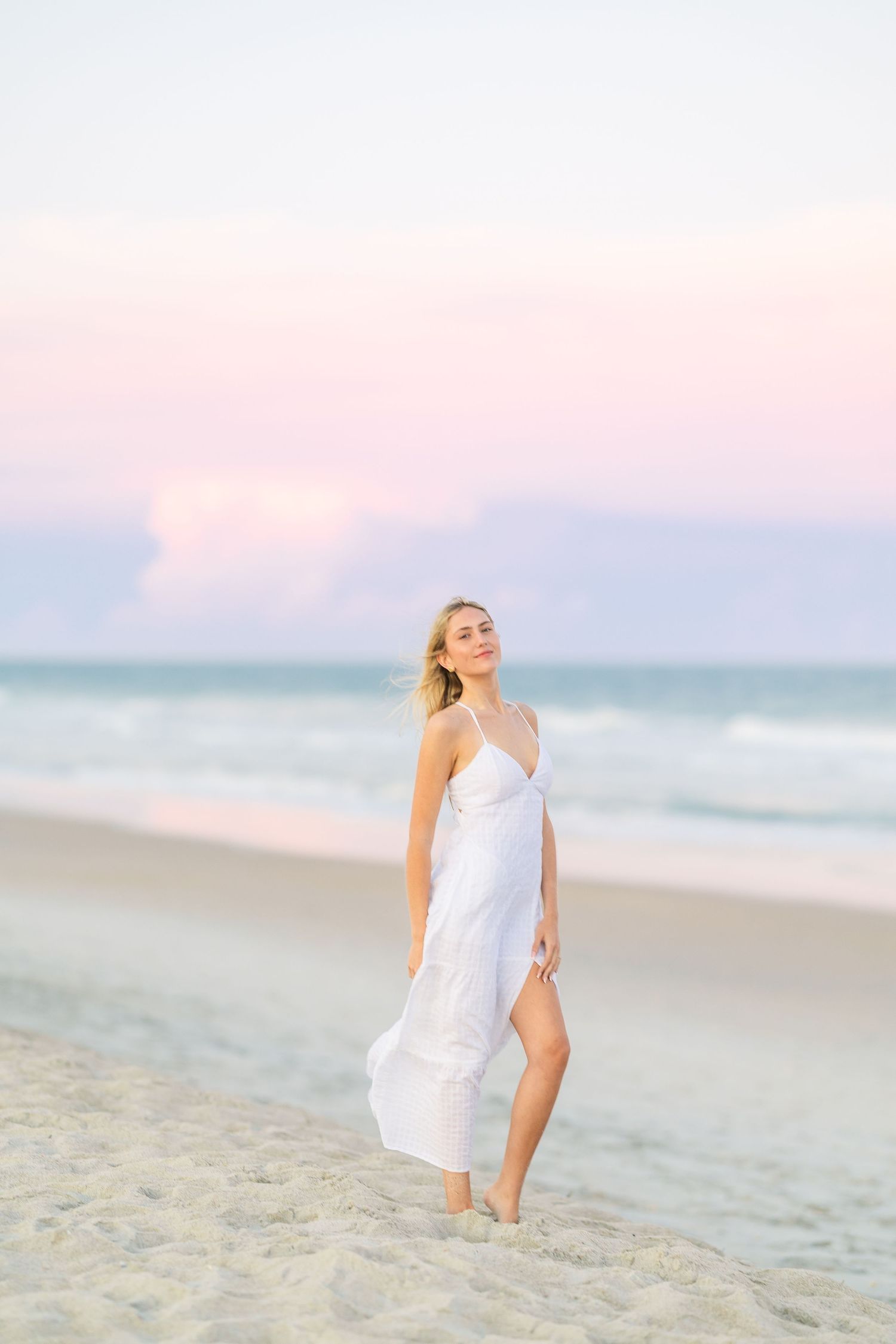 A person in a breezy white dress stands on beach sand during a pastel sunset.