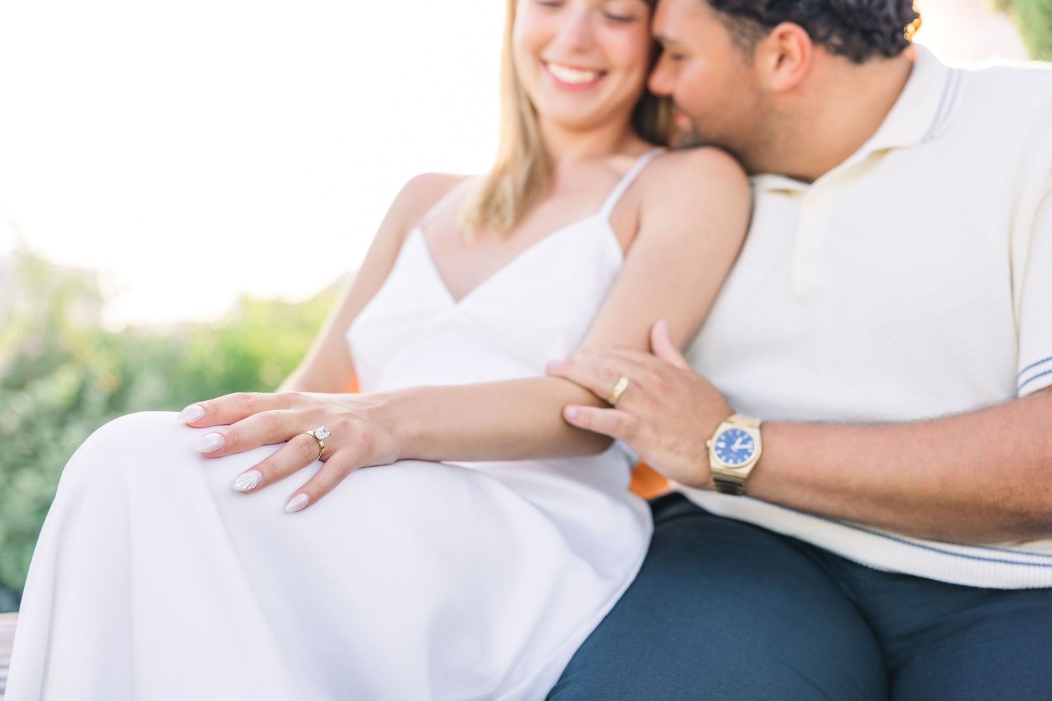 Couple sharing an intimate moment, woman displaying diamond engagement ring while man kisses her cheek outdoors.