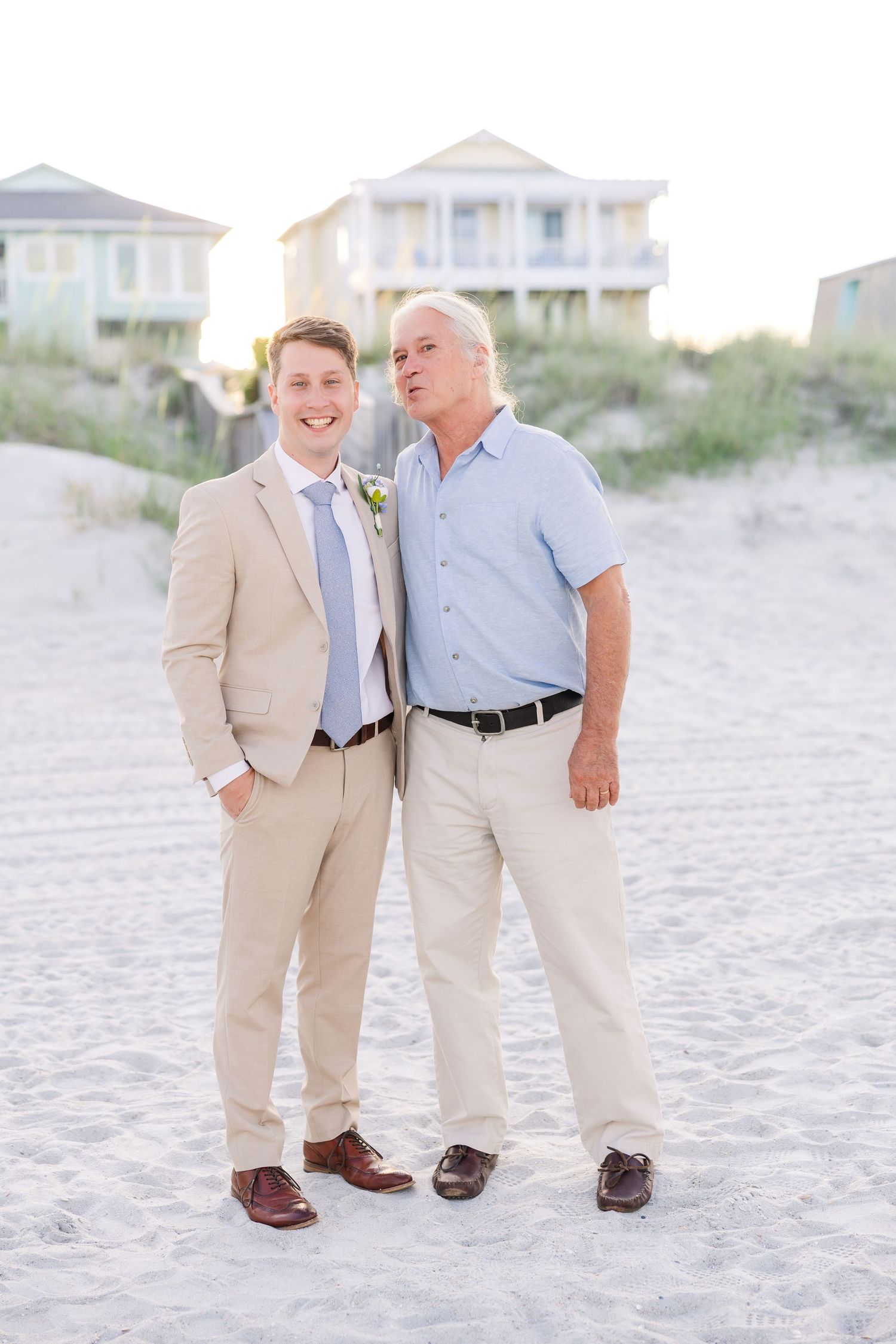 Smiling groom in tan suit poses with older man in blue shirt on sandy beach at sunset in Carolina Beach near Wilmington, NC.