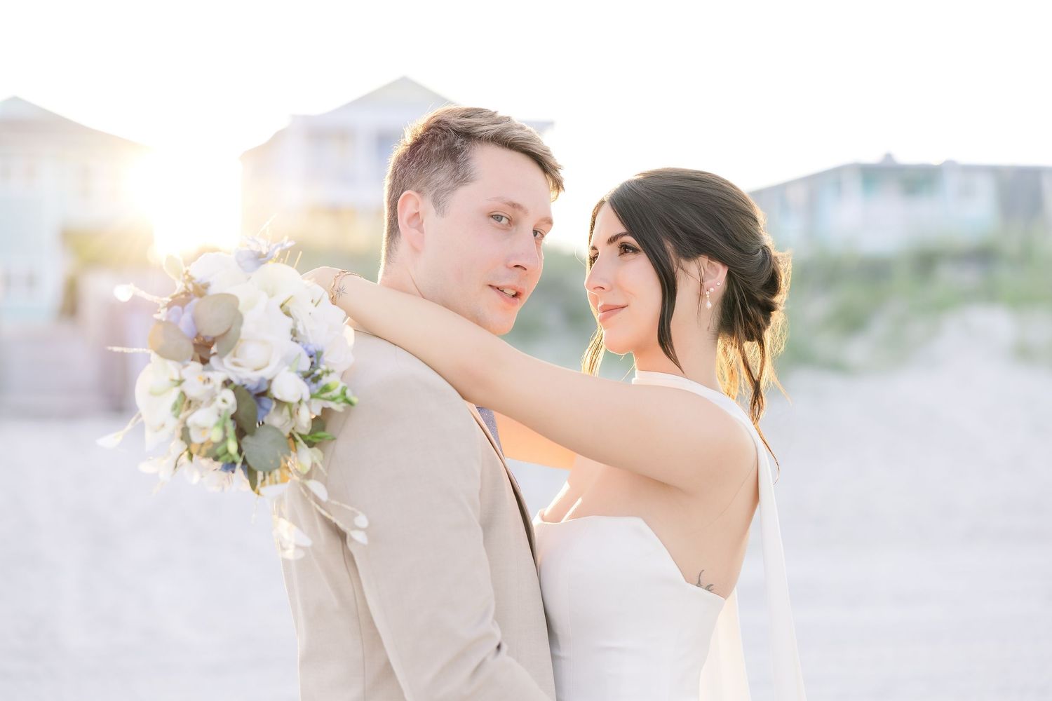 Bride and groom embrace on a sunny beach, she holds a white bouquet while wearing a strapless gown and he wears a tan suit in Carolina Beach