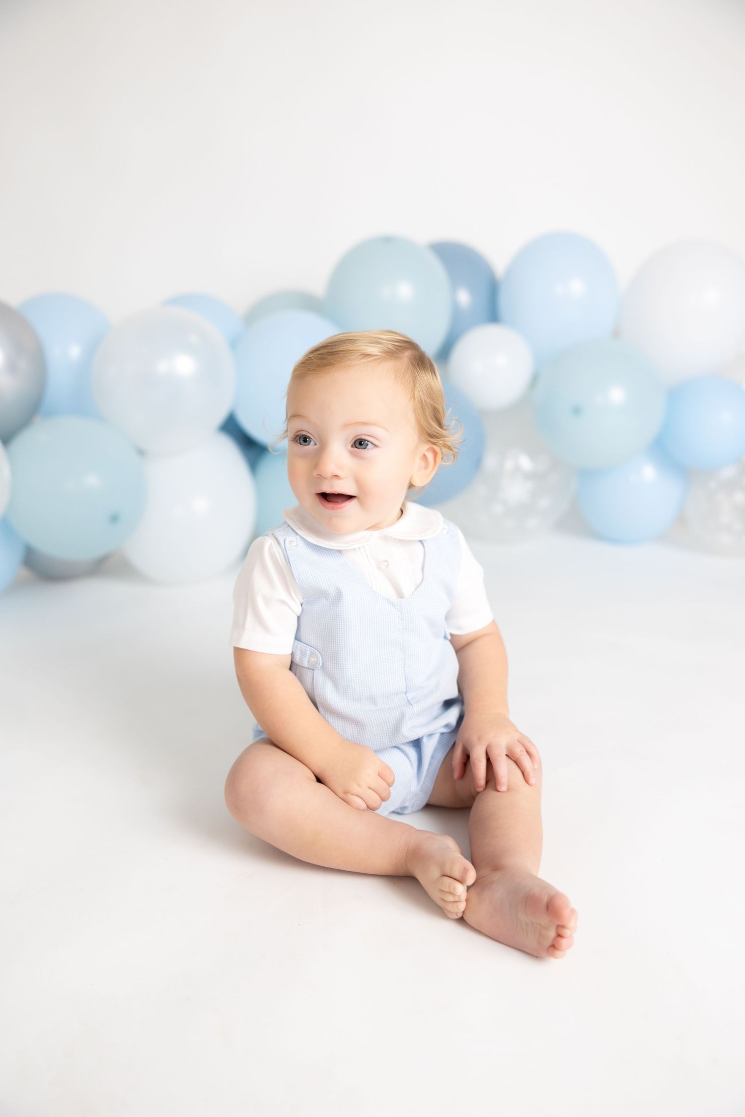 Baby in white outfit sits on white floor with blue balloon garland backdrop.