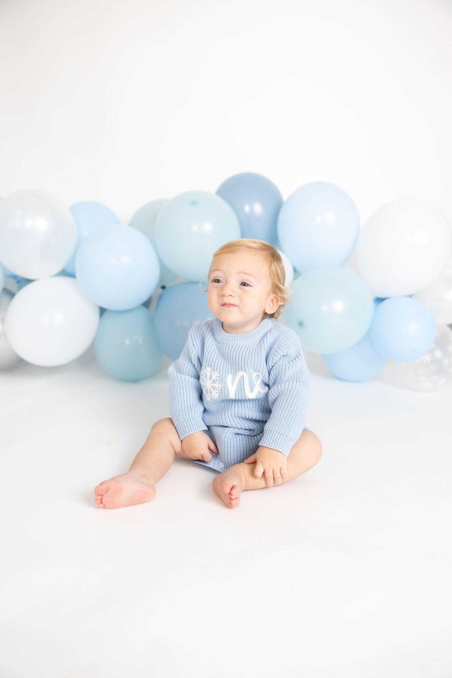 Baby in blue outfit sits on white floor with decorative blue and white balloons in background.