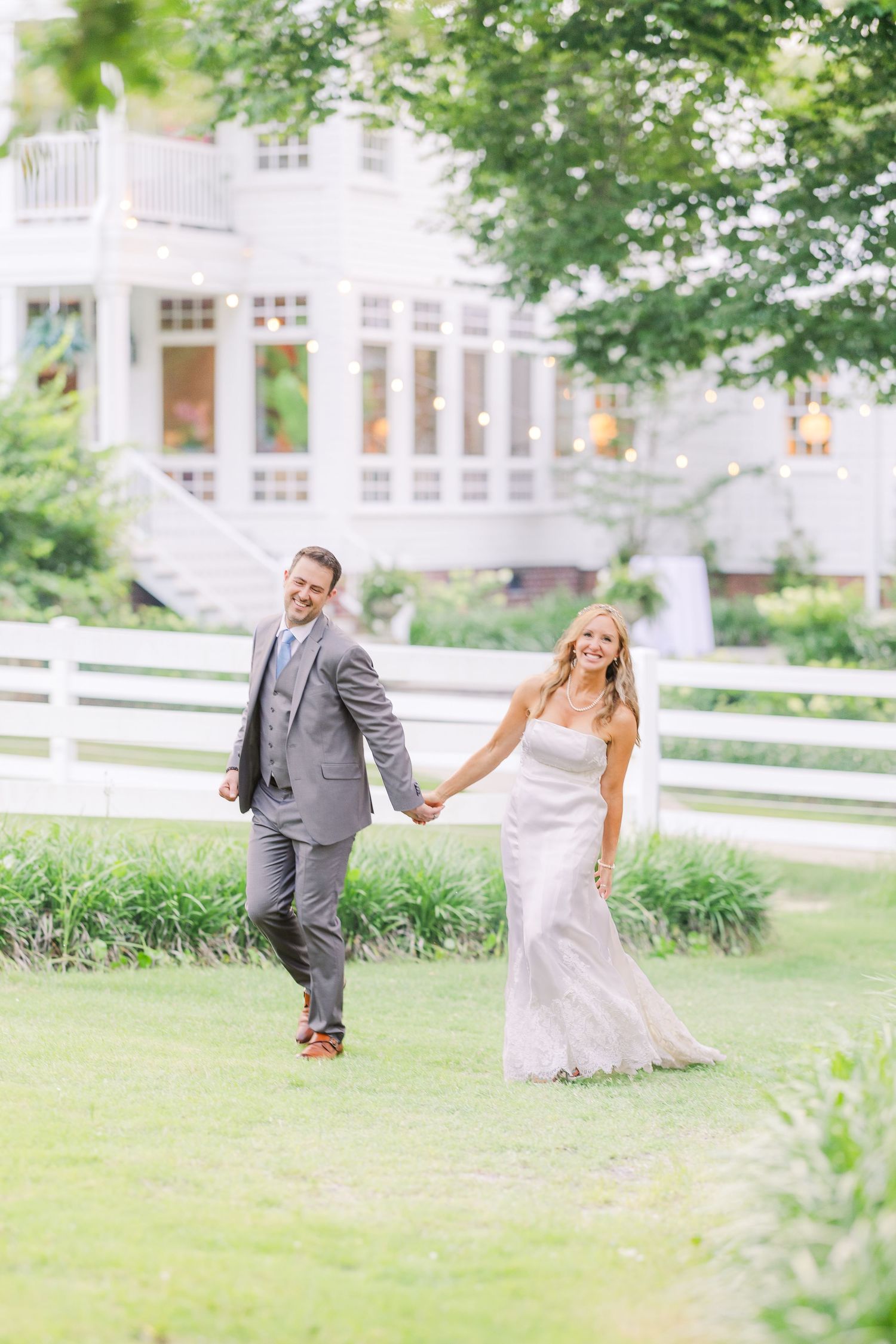 A couple in wedding attire holds hands while walking on a lush lawn with a white house in the background.