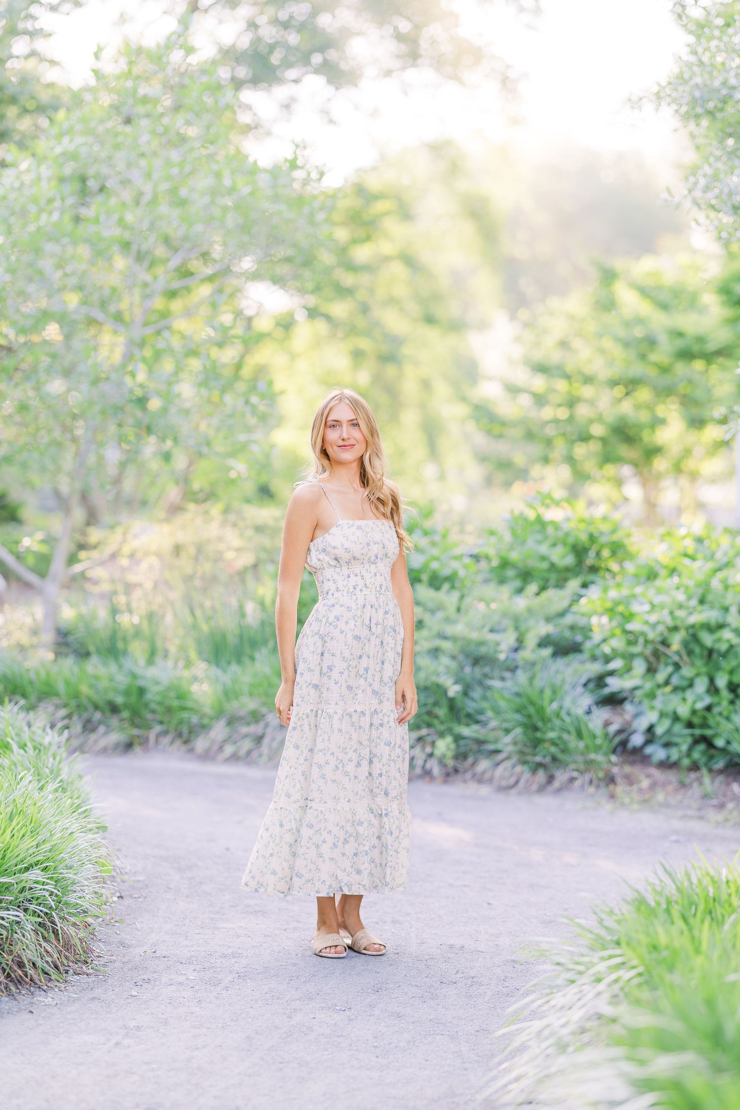 A floral sundress is worn in a sunlit garden path surrounded by lush greenery and flowers.