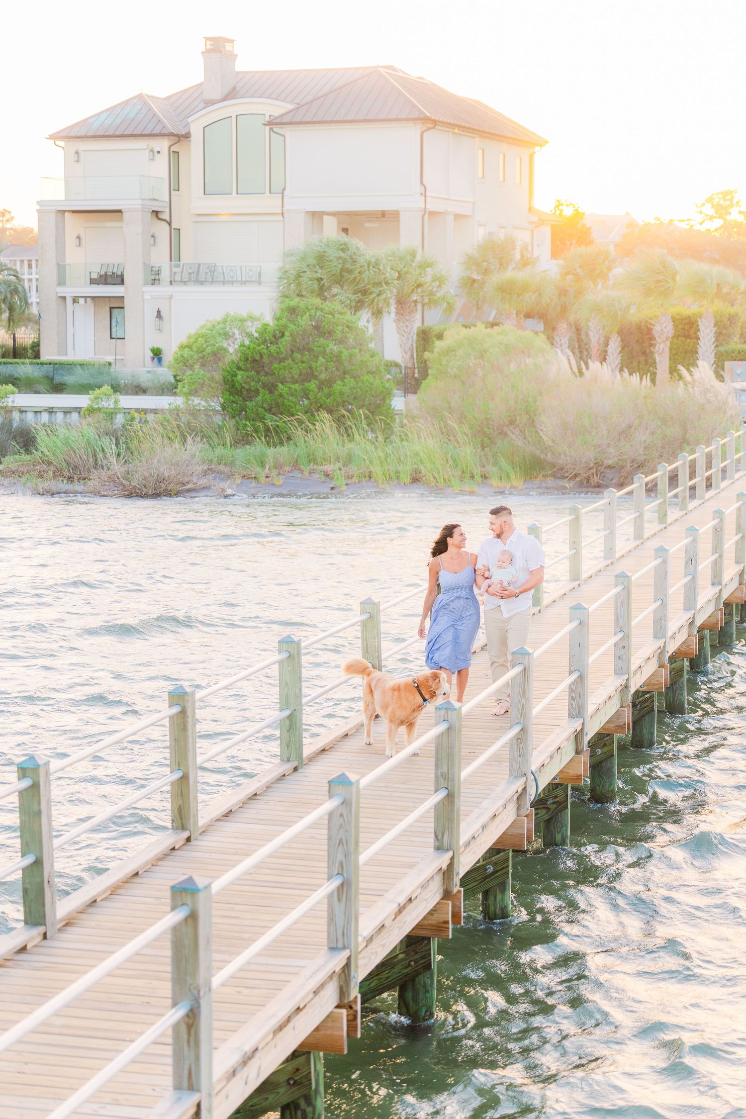 A family walks along a coastal boardwalk with their dog as the sun sets behind a beach house.
