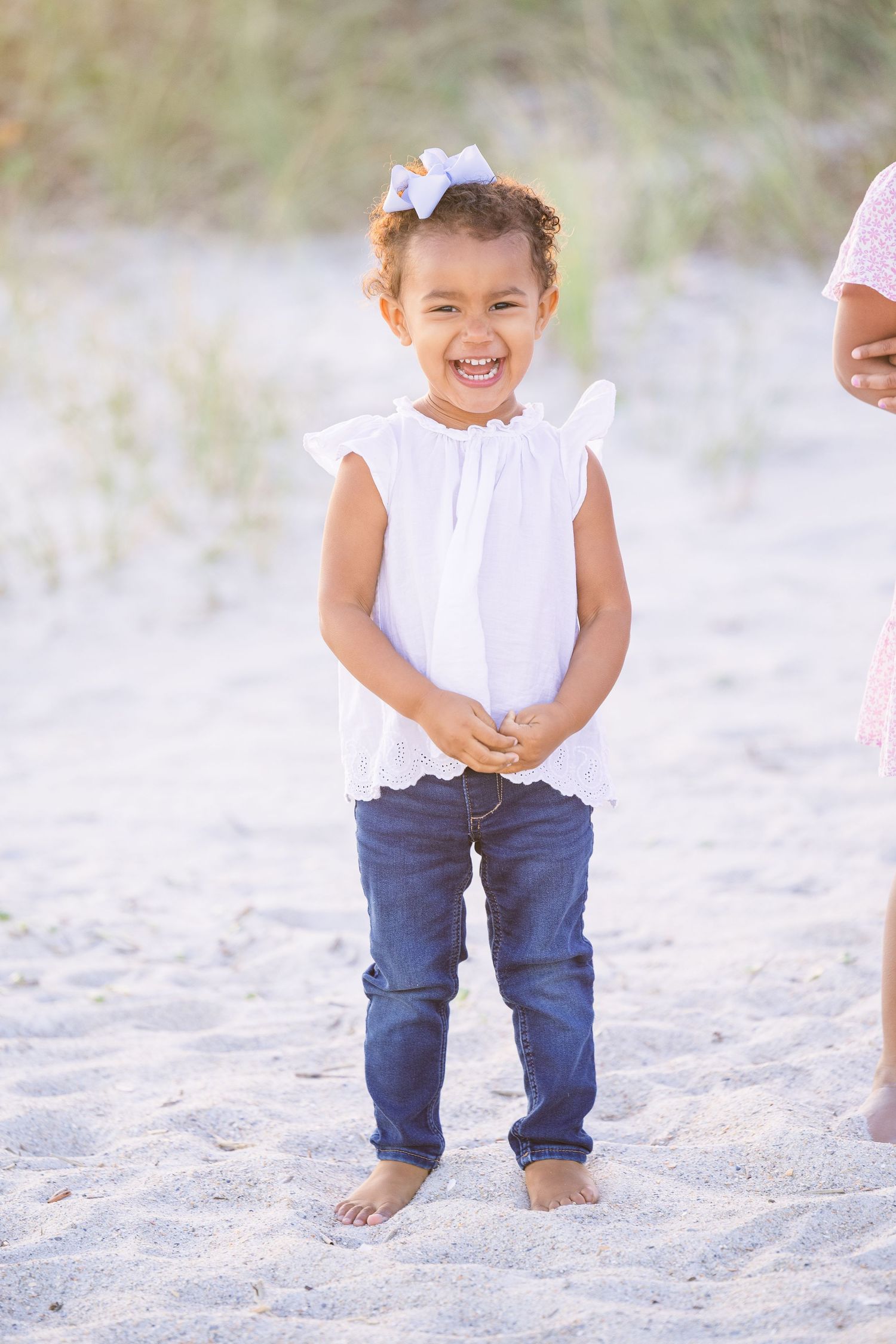 A child in a white top and jeans laughs joyfully while standing barefoot in the sand.
