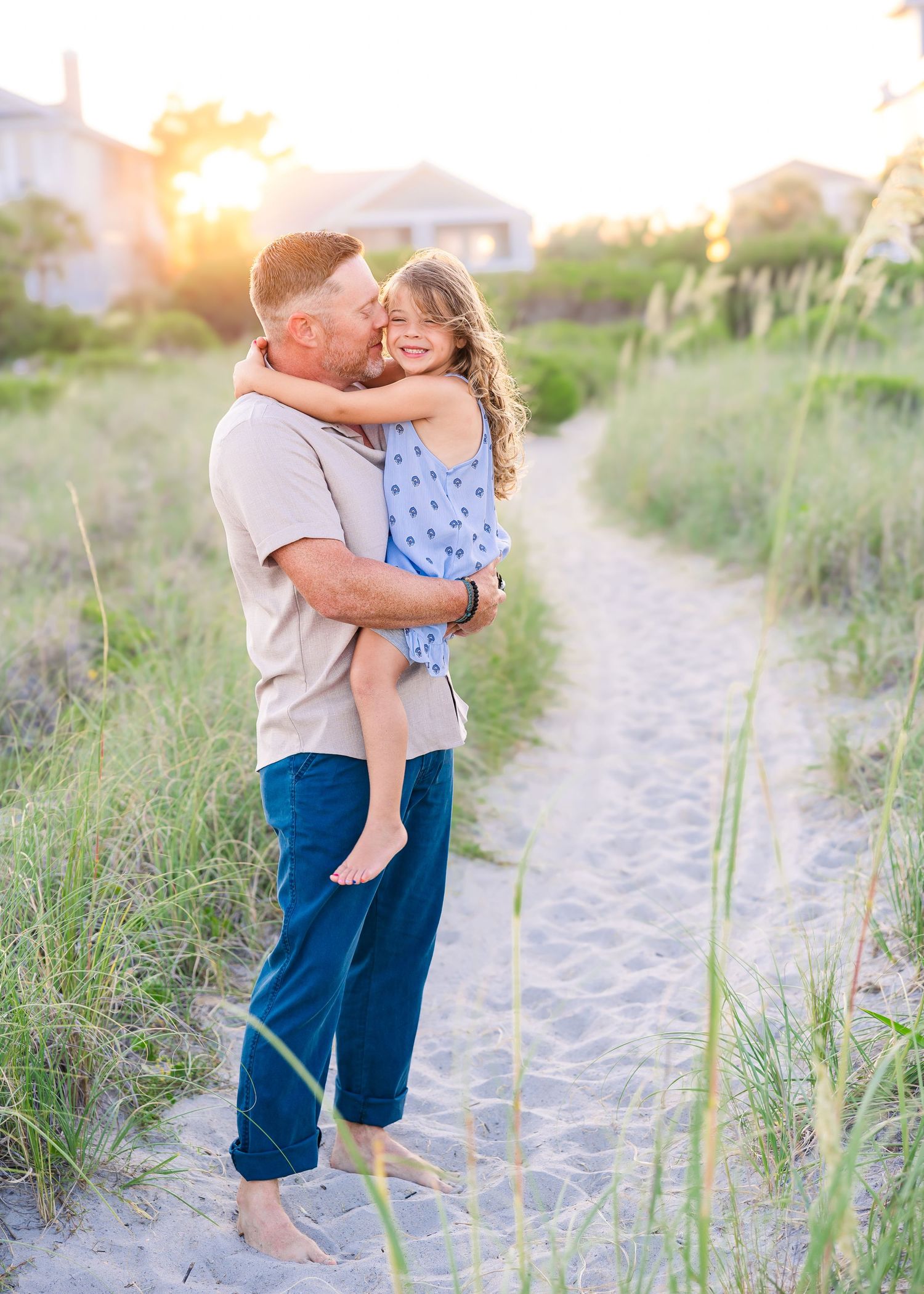 A father kisses his smiling daughter while holding her on a sandy beach path at sunset.