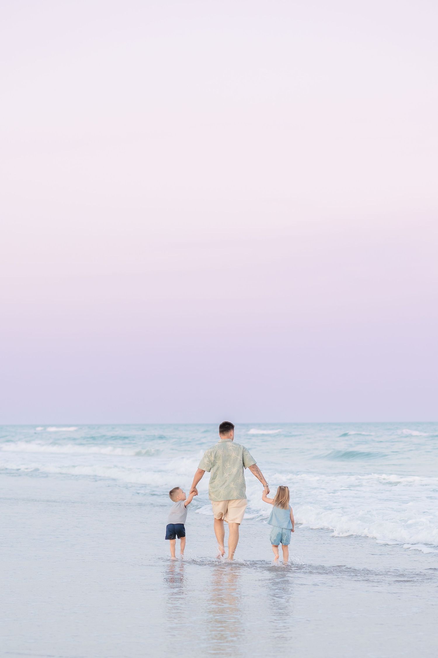 Father walking hand-in-hand with two young children toward ocean waves under a pastel pink sky.