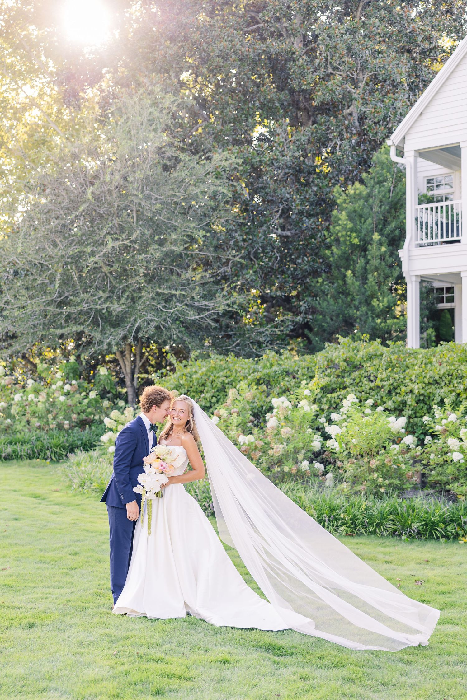 Bride and groom share a romantic kiss on lush green lawn, her cathedral veil flowing, beside a white estate.
