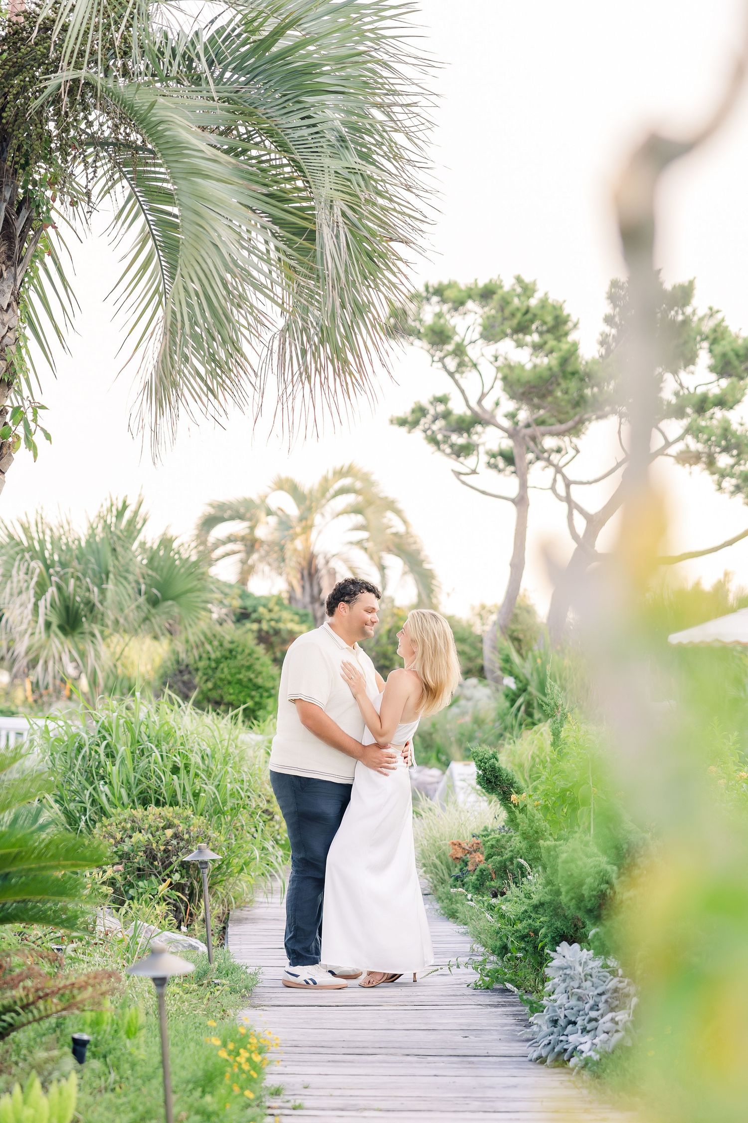 Couple embracing on a scenic wooden boardwalk lined with tropical plants and palm trees at sunset.