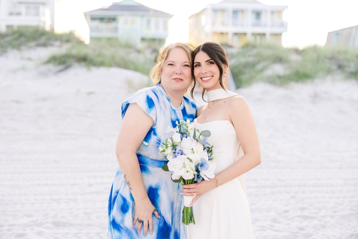 Bride in white gown holding blue and white bouquet poses with woman in blue floral dress on sandy beach in Carolina Beach near Wilmington, N