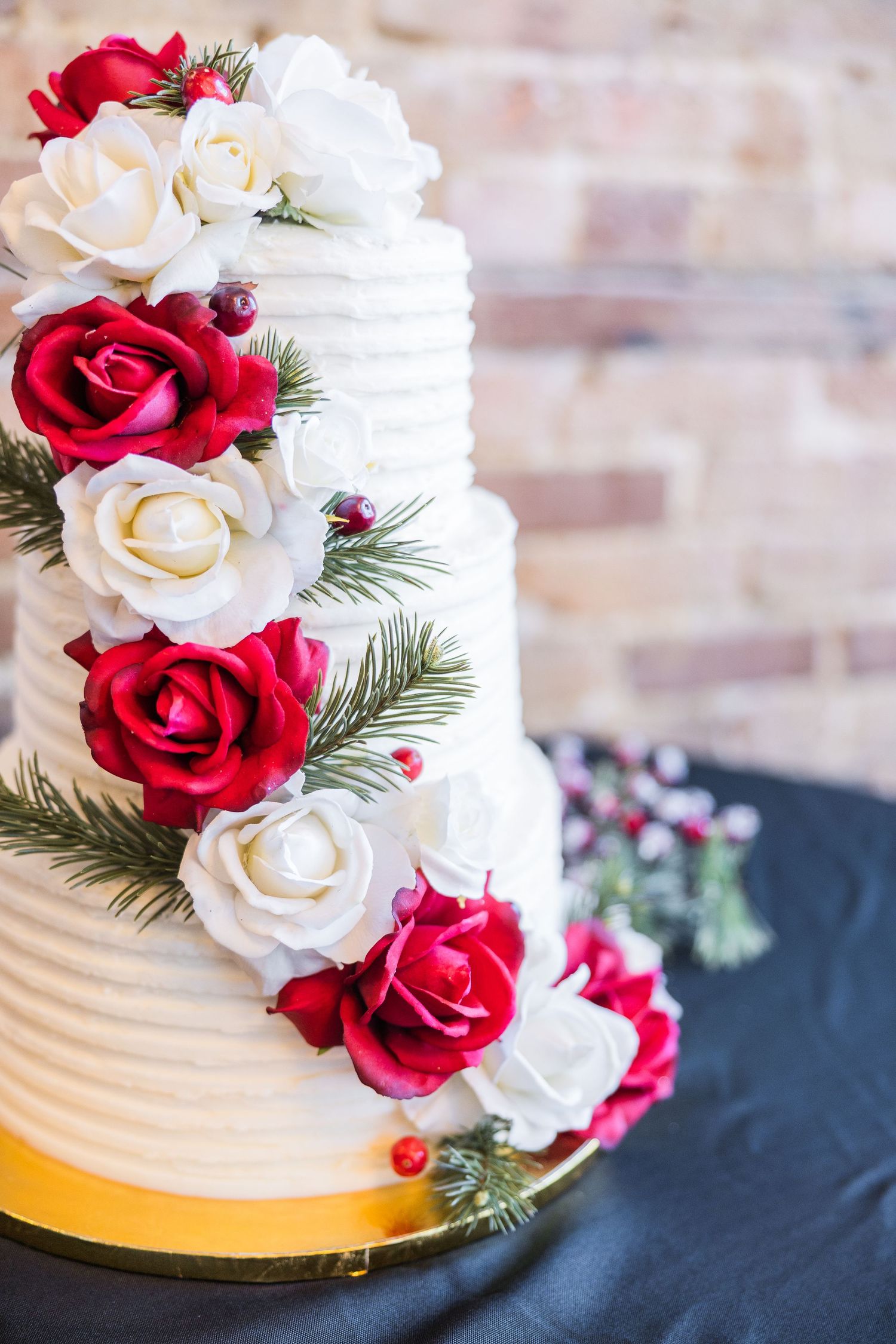 A white wedding cake decorated with red and white roses and evergreen sprigs on a navy tablecloth.