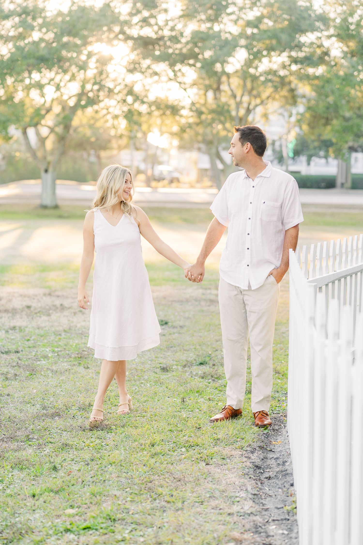 Couple holding hands while walking through a grassy area with white fence in golden sunlight.