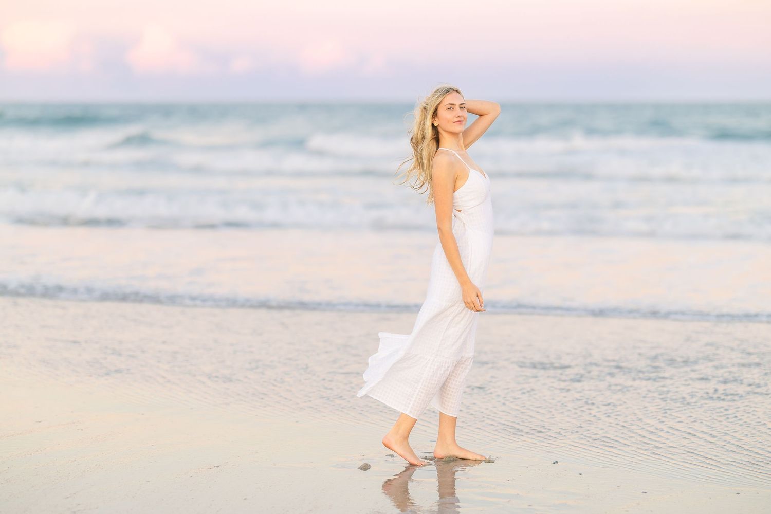 Woman dances joyfully on beach wearing white summer dress at sunset.