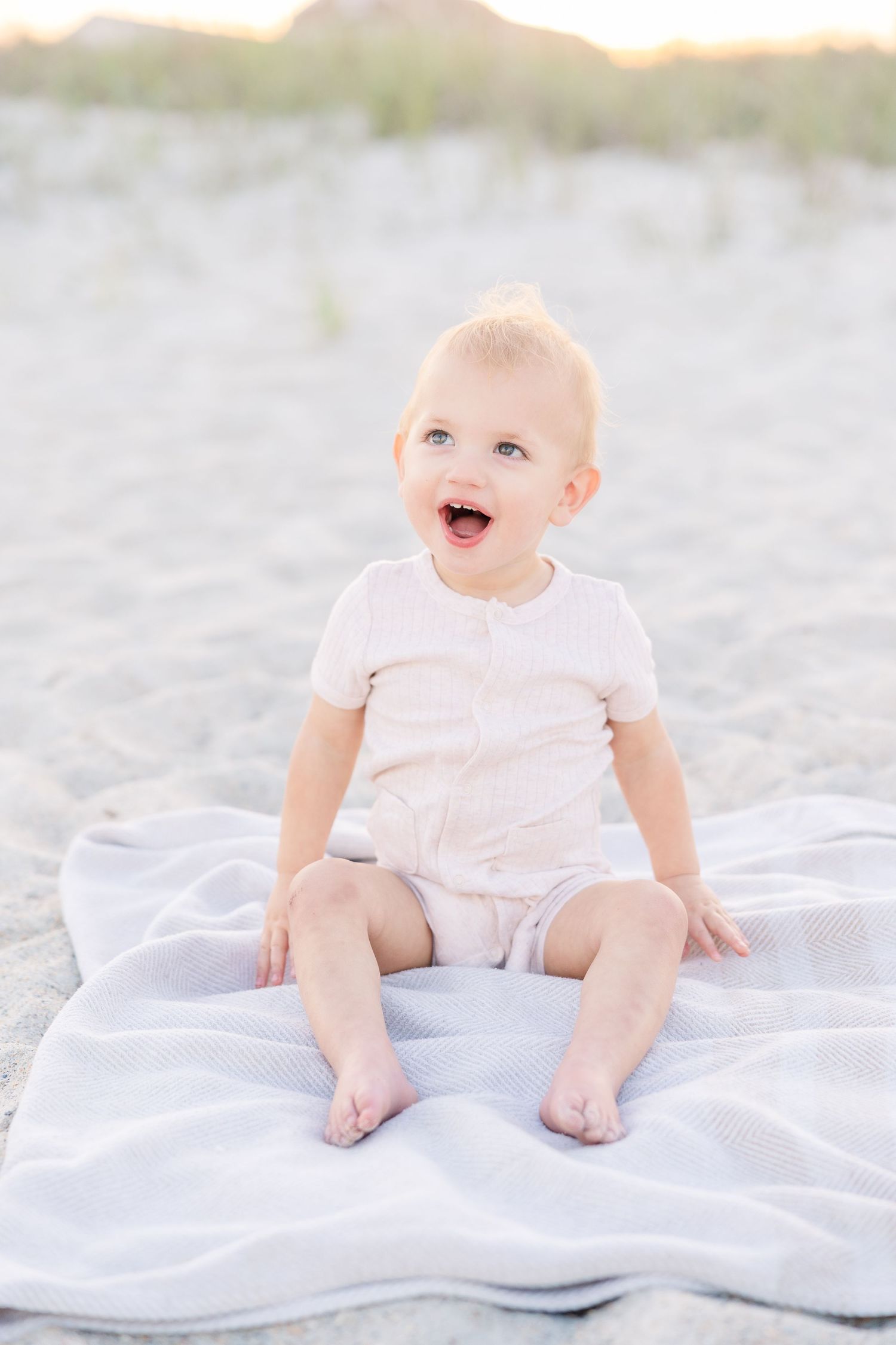 Happy baby sitting on white blanket on sandy beach, wearing light pink romper, laughing with joy.