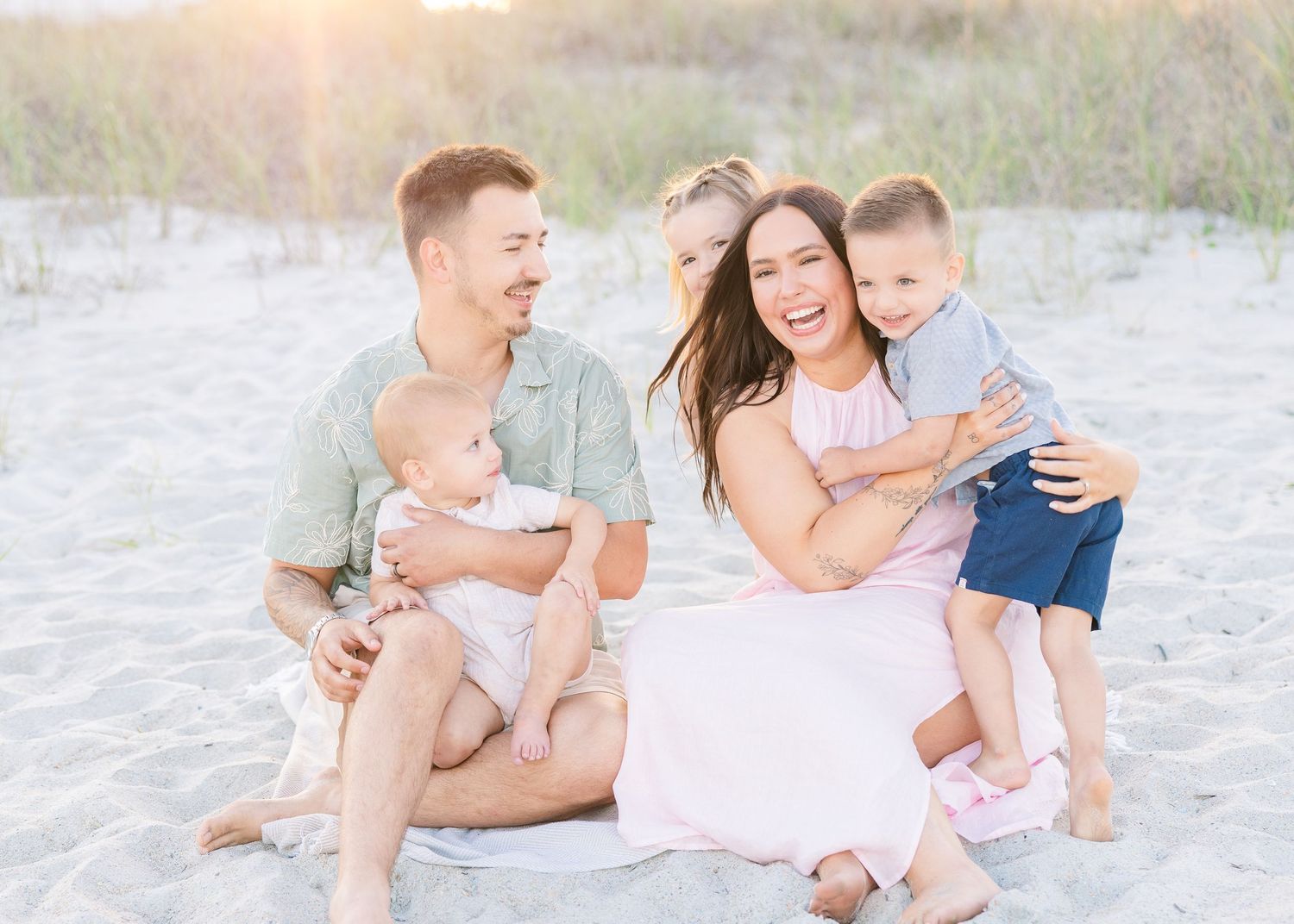A laughing family of five sits together on a sandy beach blanket in warm golden light.