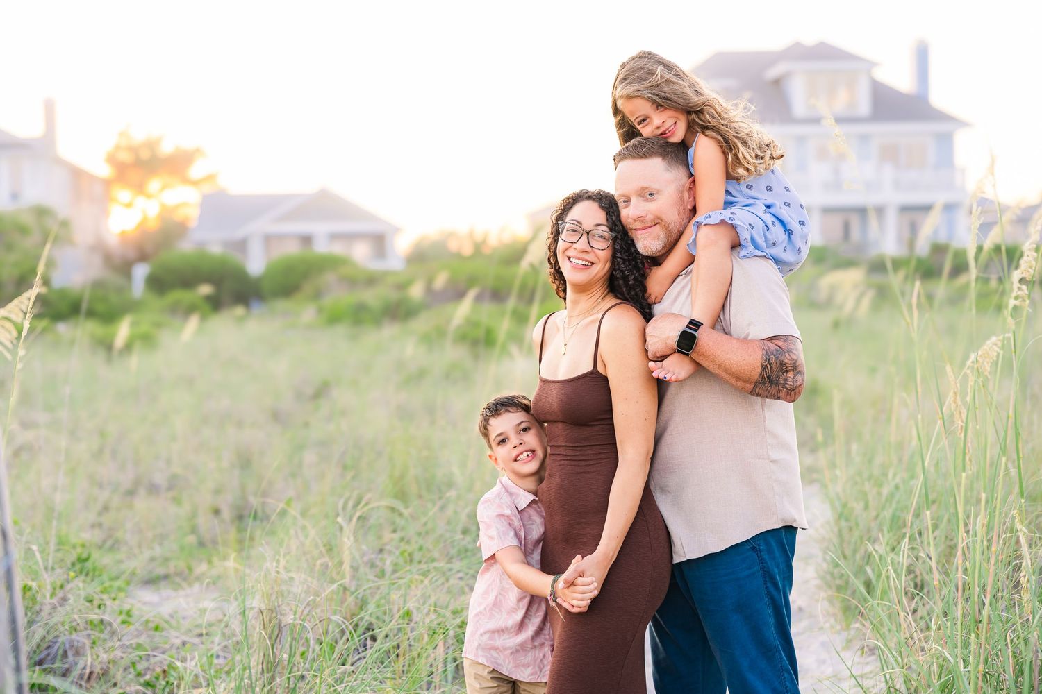 Family of four hugging and smiling together in a coastal grass field at golden hour.