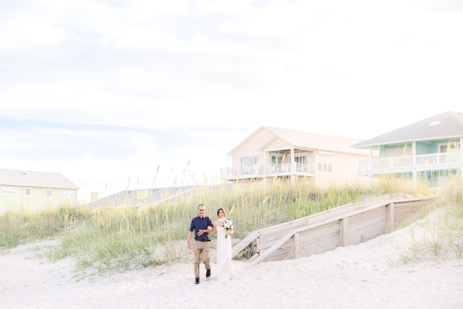 Couple in wedding attire walking hand in hand on a sandy beach with coastal homes in the background in Carolina Beach near Wilmington, NC.