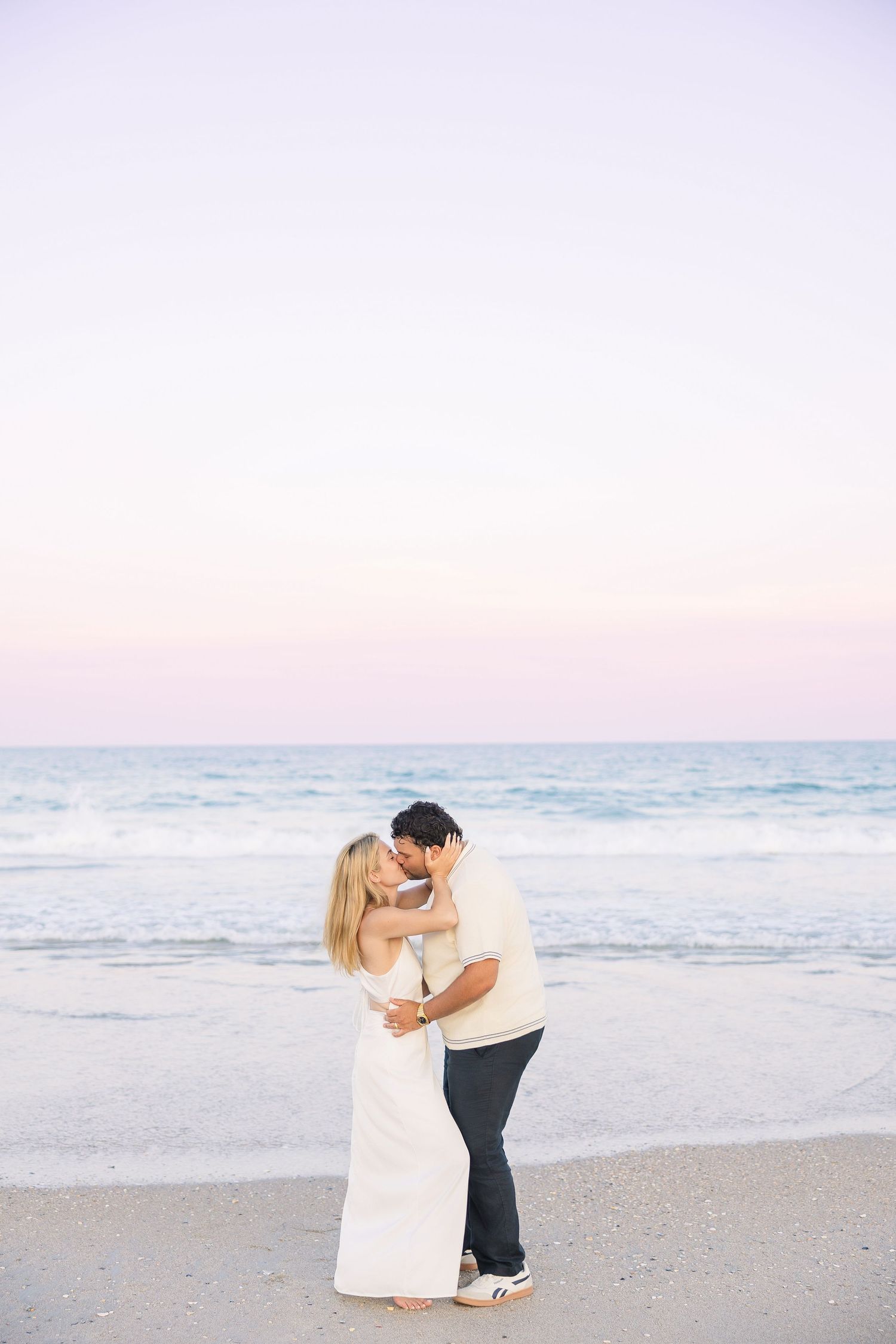 Couple embracing on sandy beach at sunset with pastel purple sky and ocean waves behind them.