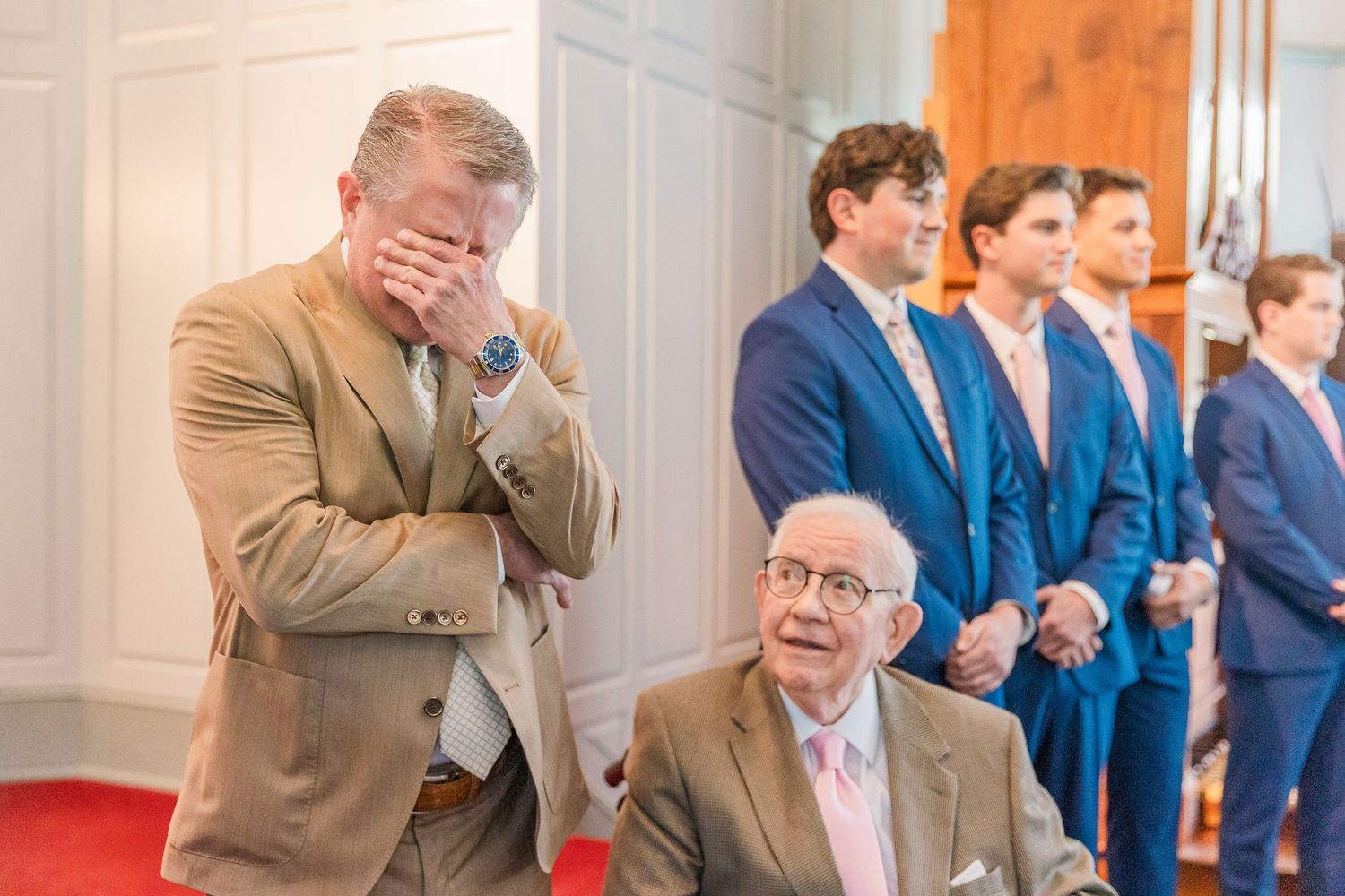 Groomsmen in navy and tan suits line up during wedding ceremony in elegant white venue.