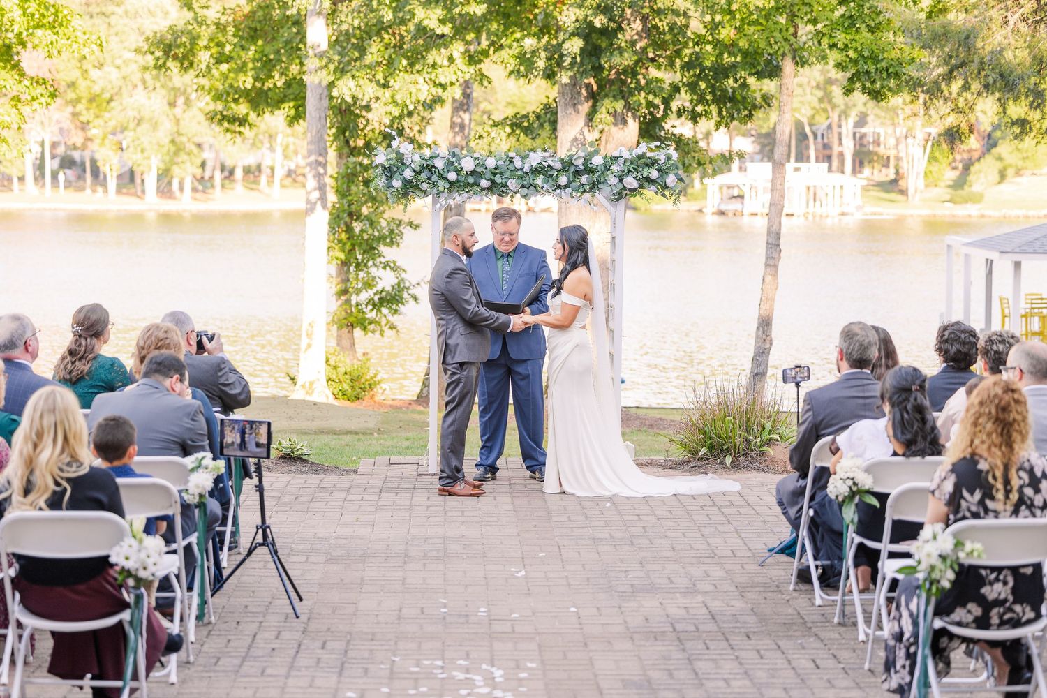 Wide shot of outdoor wedding ceremony with guests seated in rows of white chairs wedding groom bride lake Gaston photographer light airy