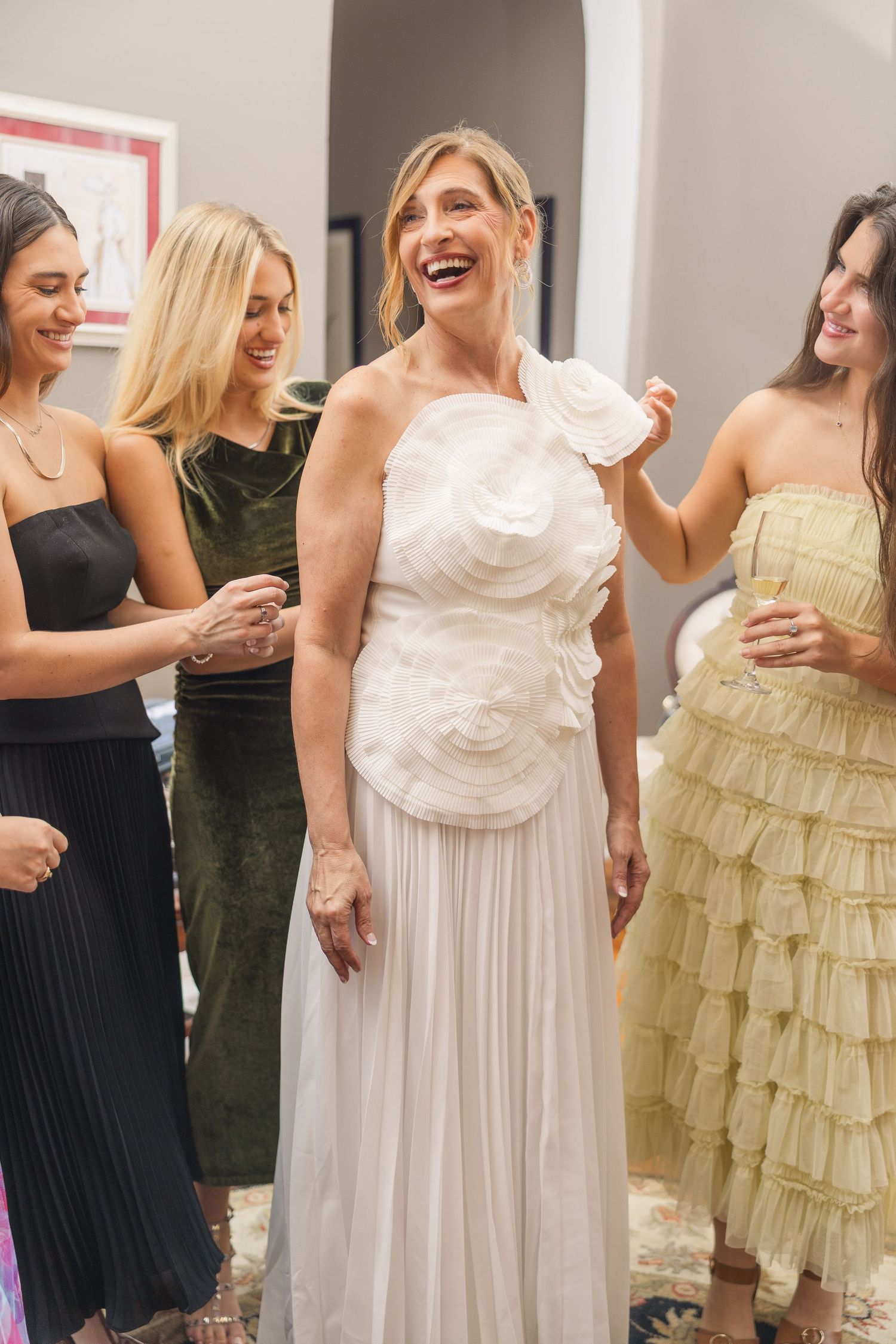 Wedding party members in white and neutral colored dresses gather together in a bright indoor space.