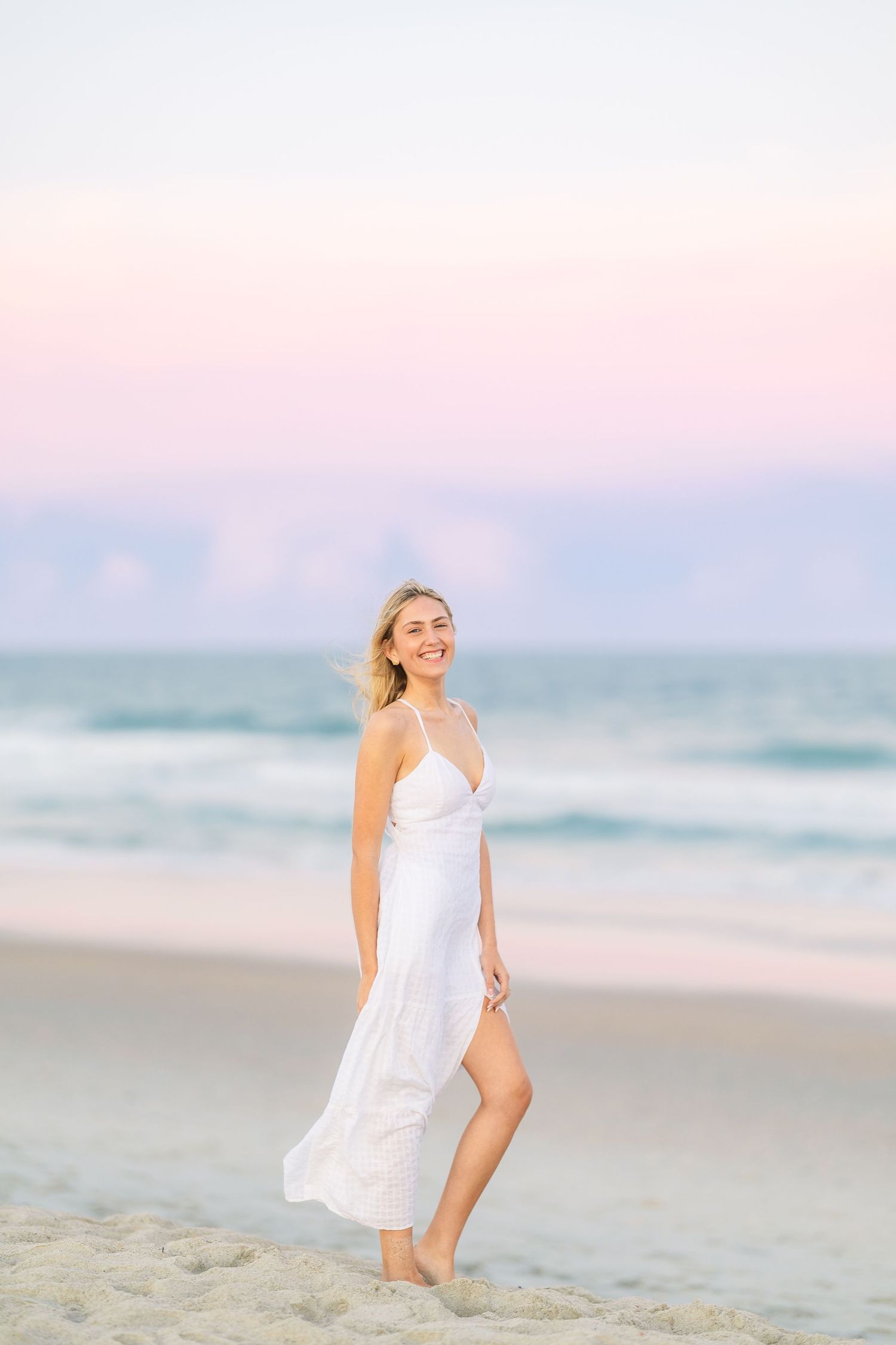 A person in a flowing white dress poses elegantly on the beach during golden hour.