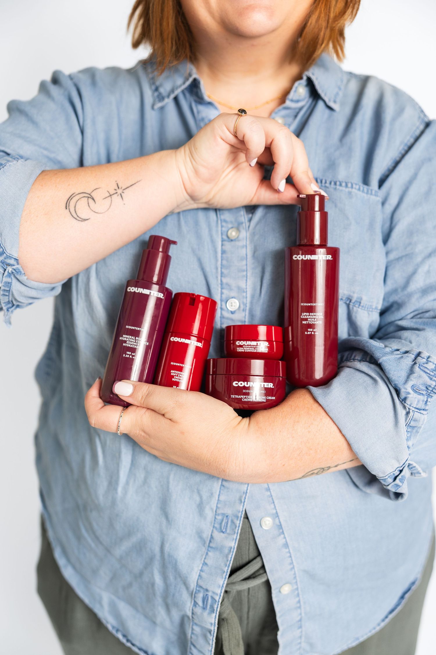 Model wearing denim shirt displays red beauty product collection in casual poses.