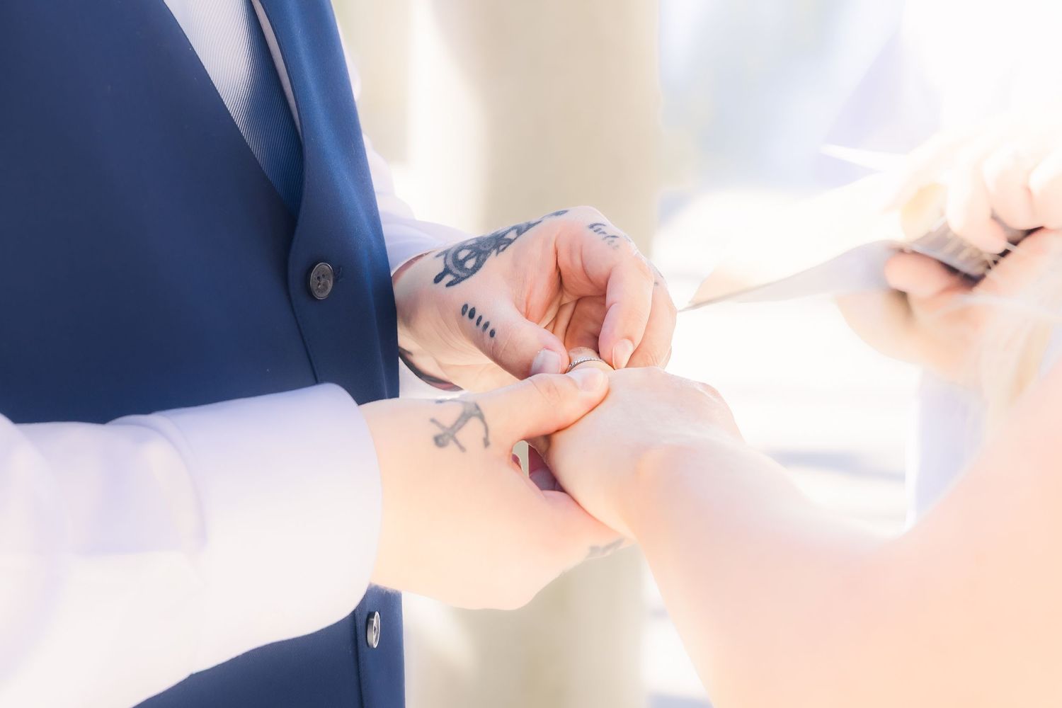 Groom in blue suit placing wedding ring on bride's finger during ceremony, with visible tattoos on hand in Airlie Gardens in Wilmington, NC