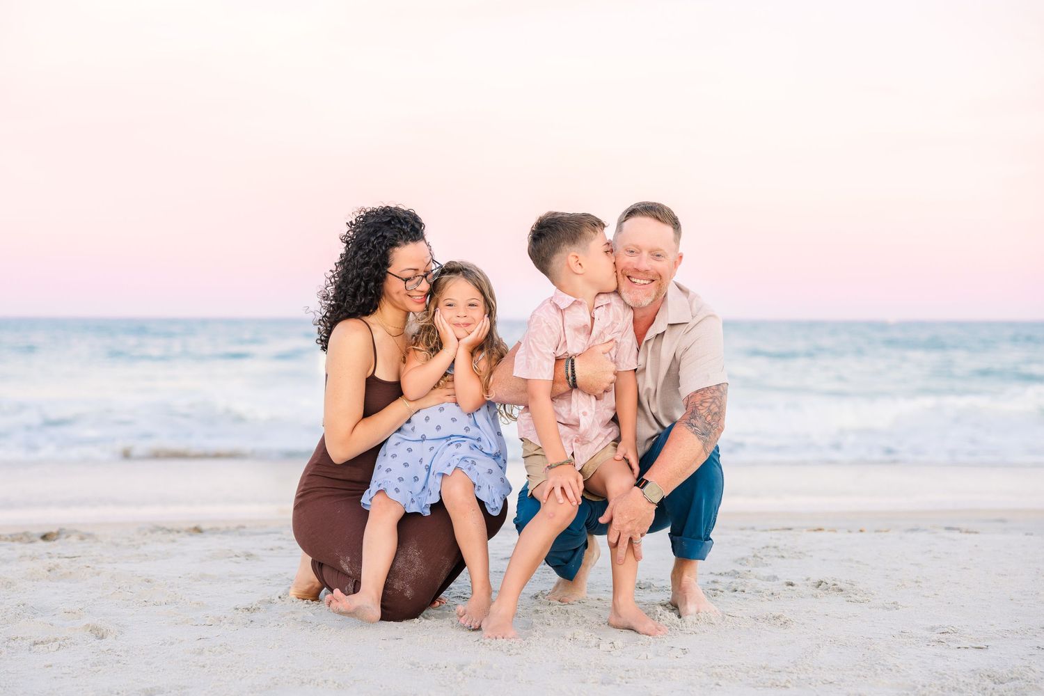 Family of four posing on sandy beach at sunset, parents crouching with two young children smiling and sharing a kiss.