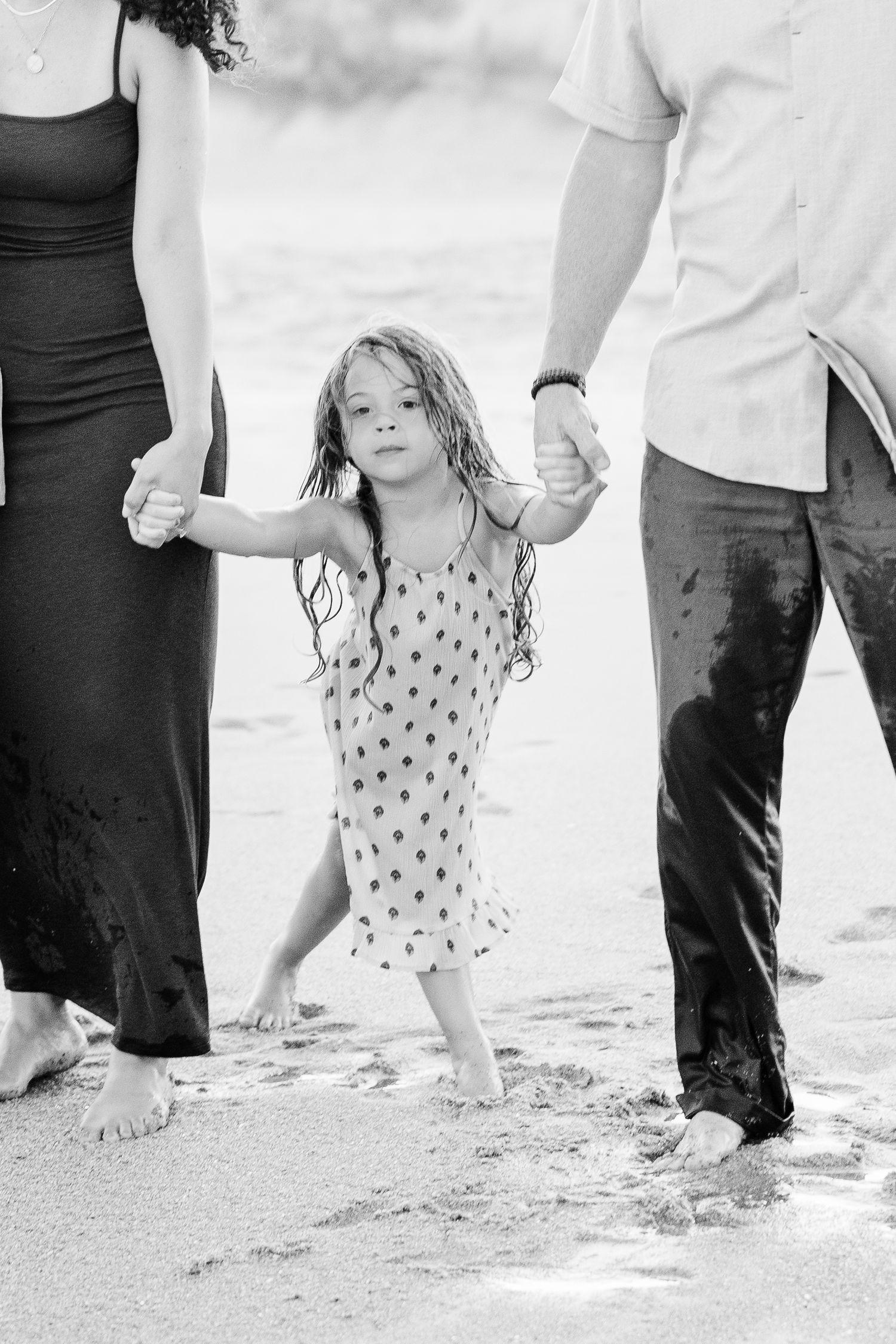 Black and white photo of a young girl holding hands with her parents while walking on the beach.