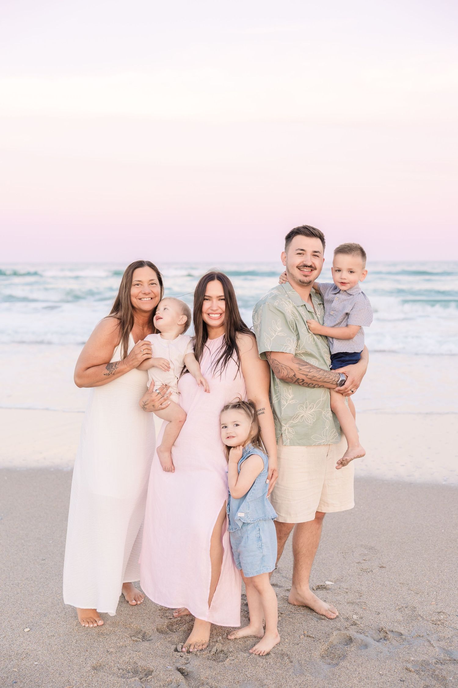 Extended family of six poses on beach at dusk, adults holding young children with ocean backdrop.
