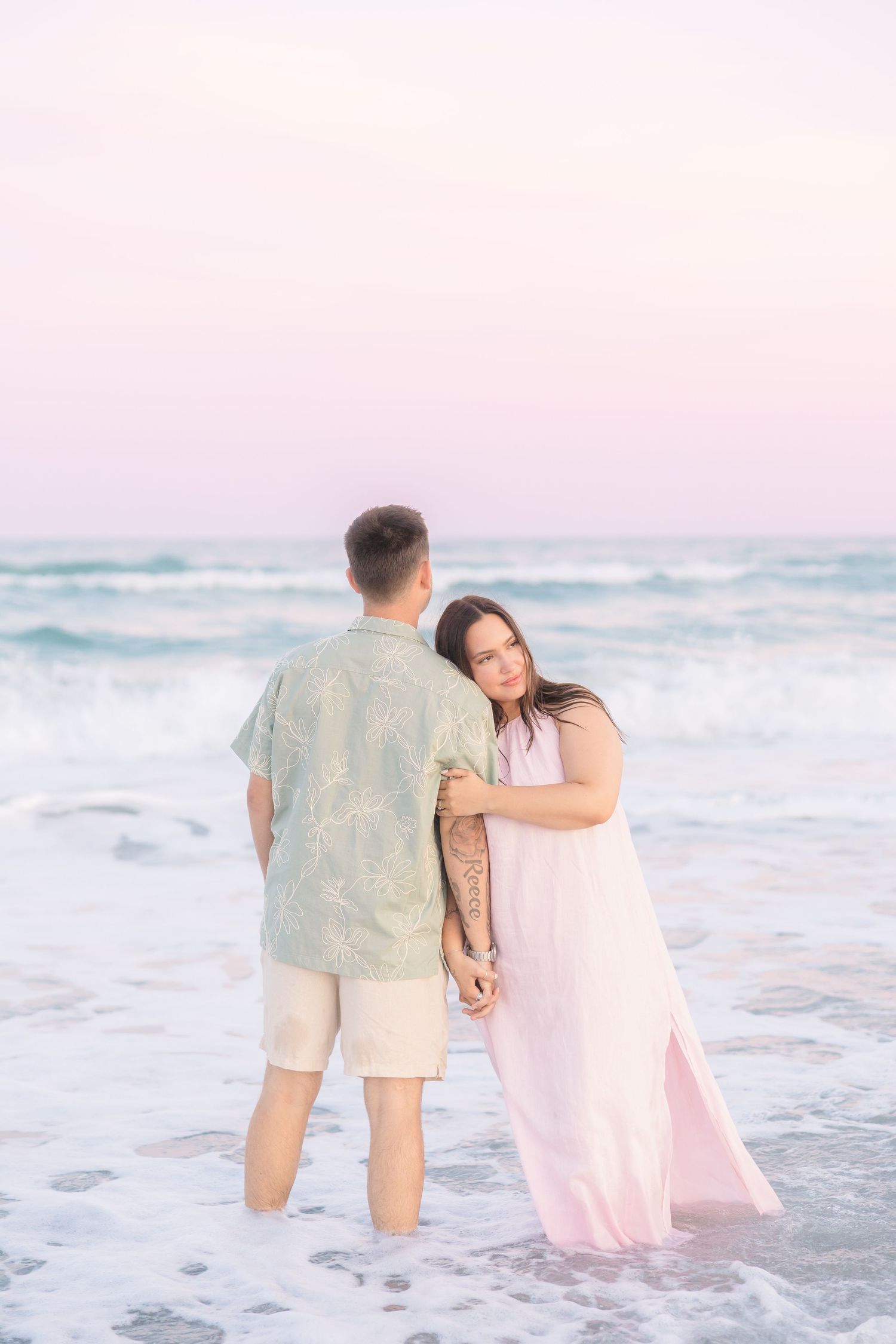 Couple standing together in ocean waves at sunset, woman in pink dress leaning on man in floral shirt.