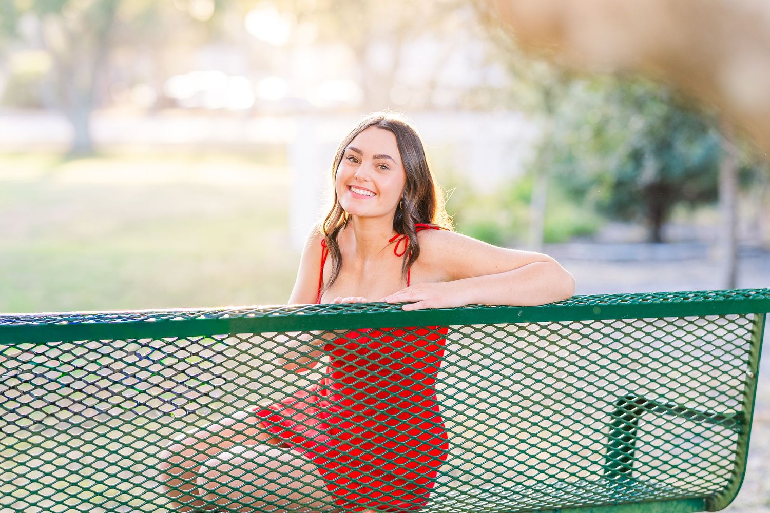 Red dress portrait leaning on green metal bench in outdoor setting.