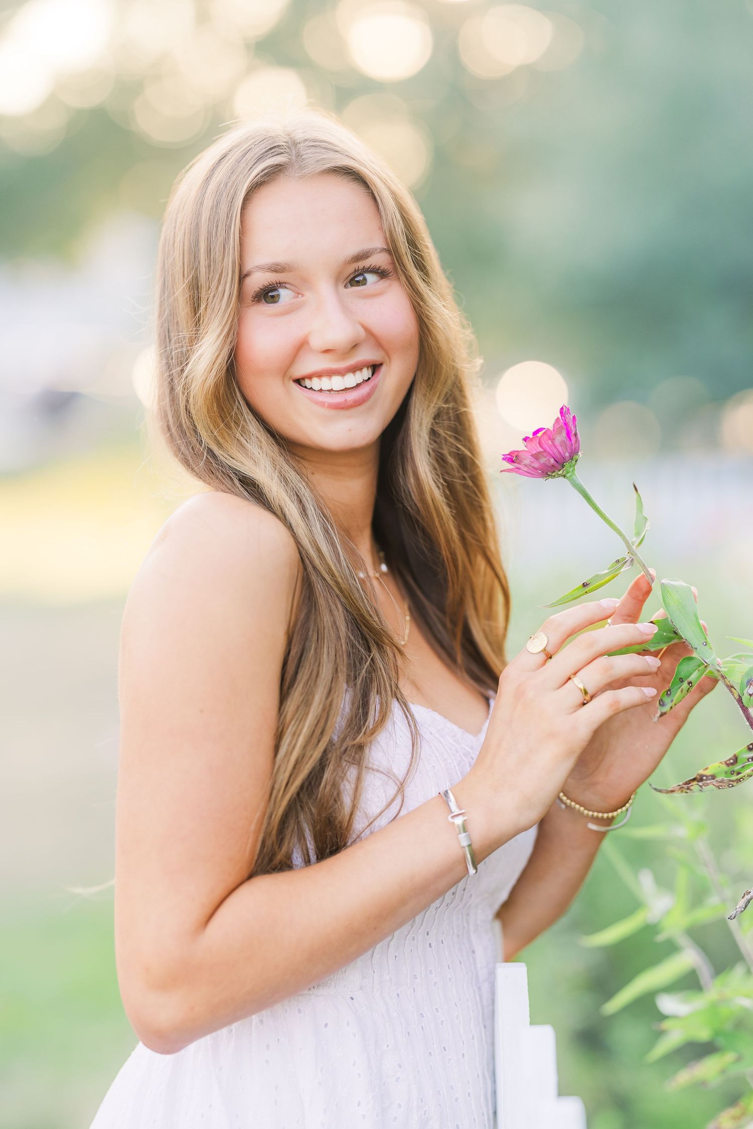 Someone in white dress holds pink flower while posing in garden with bokeh background.