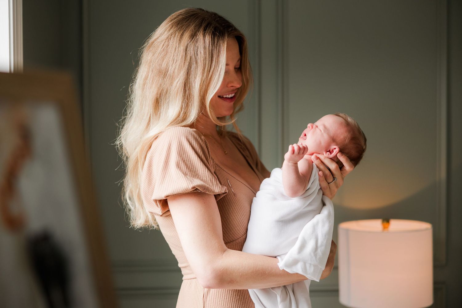 Woman in white dress holding baby in sunlit room with soft decor.