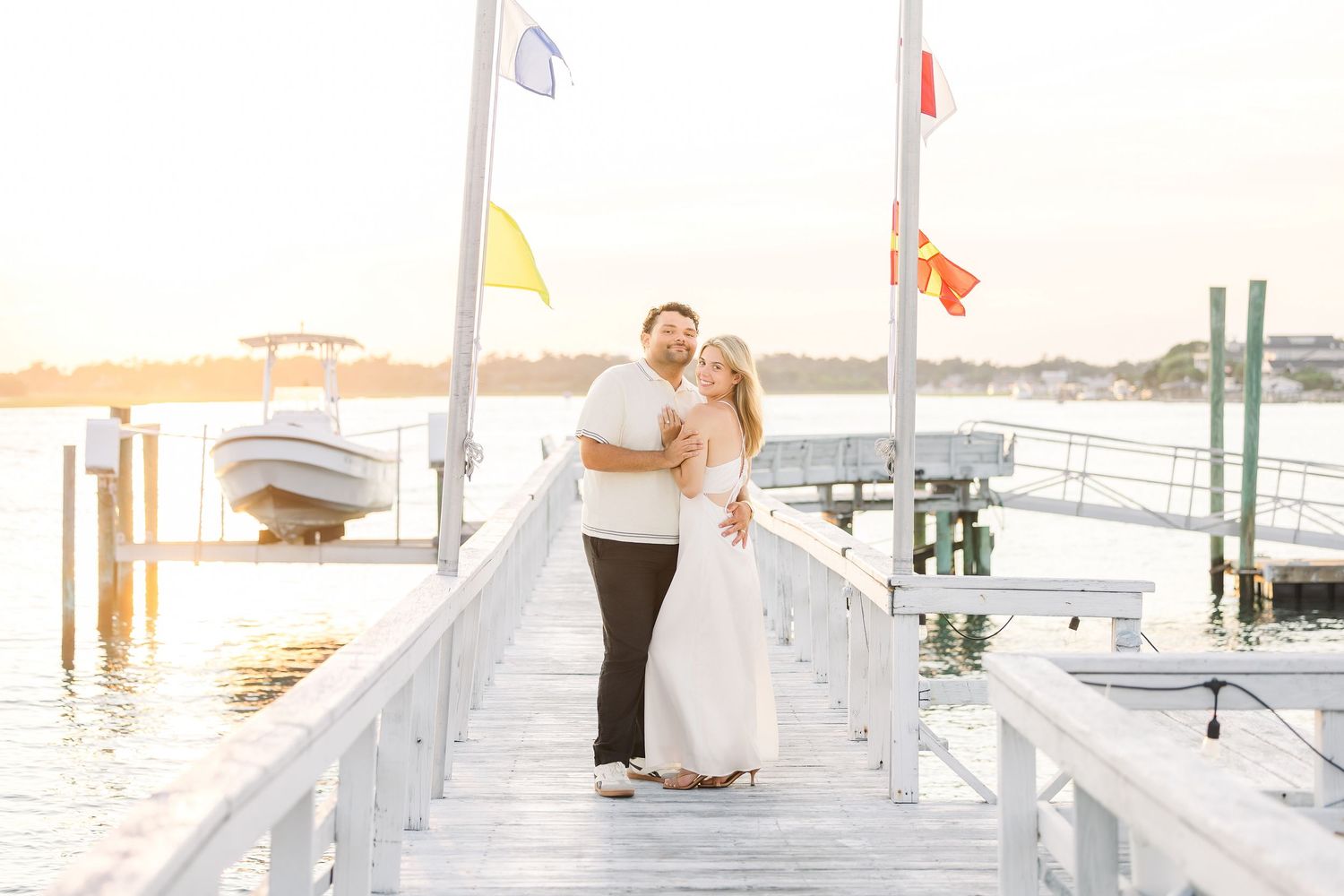 Happy couple embracing on a wooden dock at sunset with colorful nautical flags and boats in the background.
